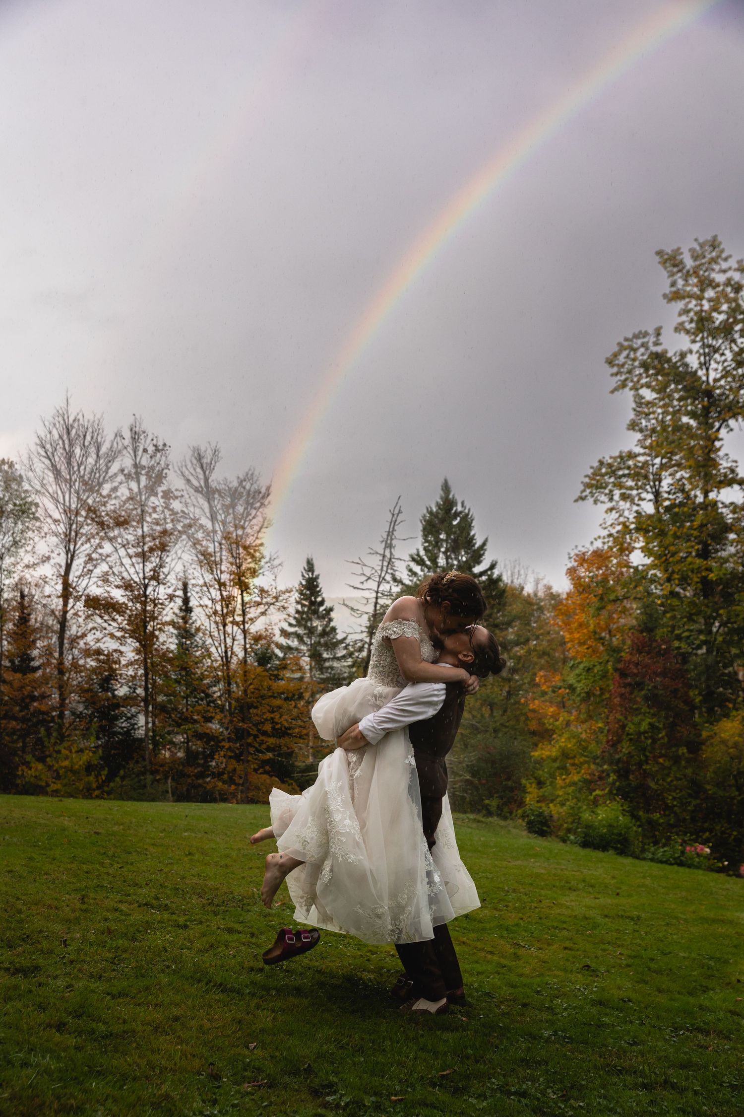 Groom lifts bride in a romantic embrace beneath a stunning double rainbow in autumn.