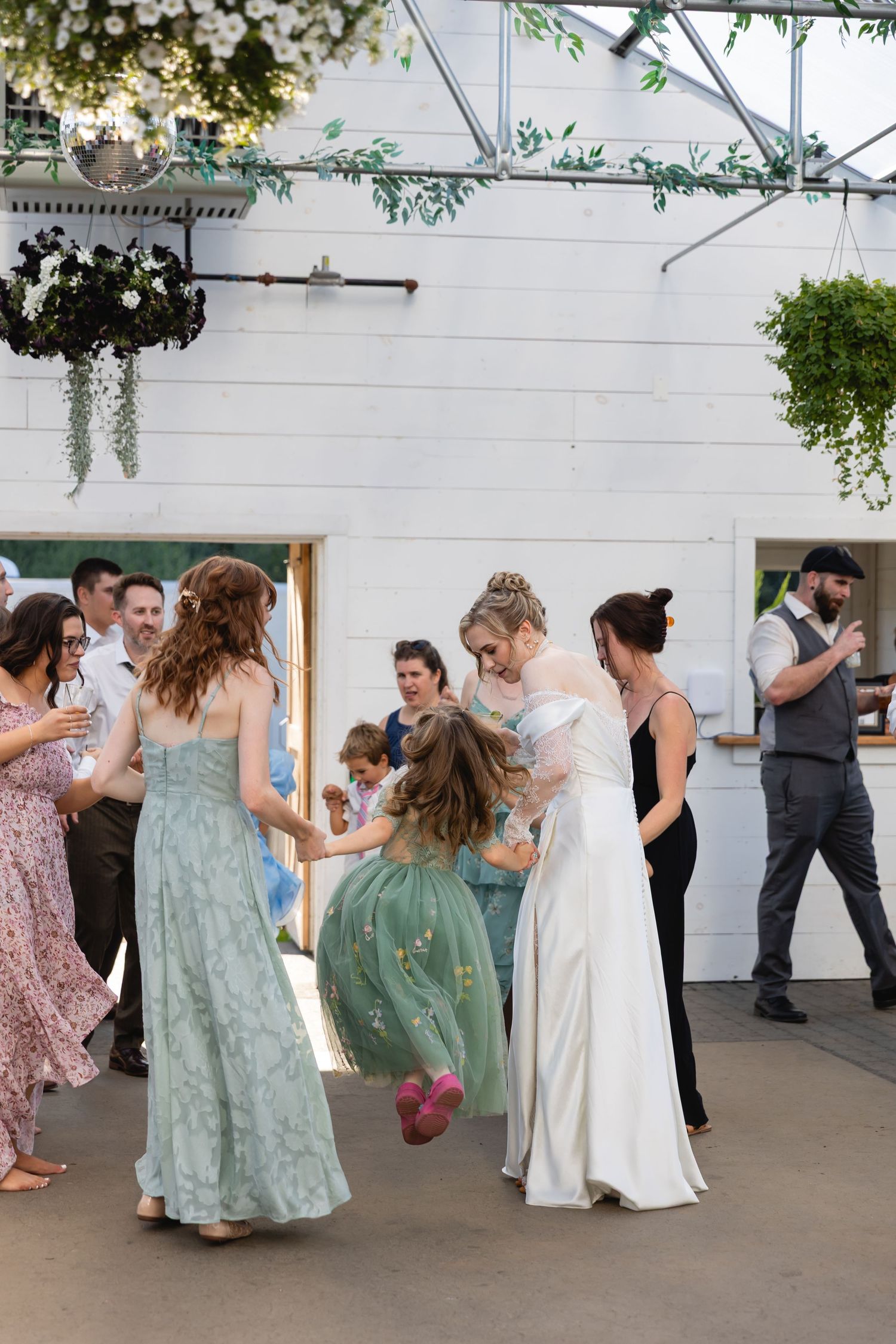 Wedding party guests dancing together in a line on an outdoor patio with greenery decorations overhead.