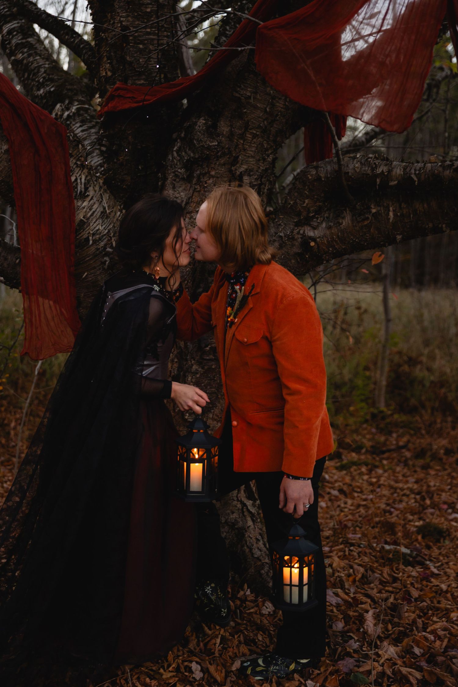 Two people share a romantic moment under red fabric draped between trees in a moody autumn forest setting.