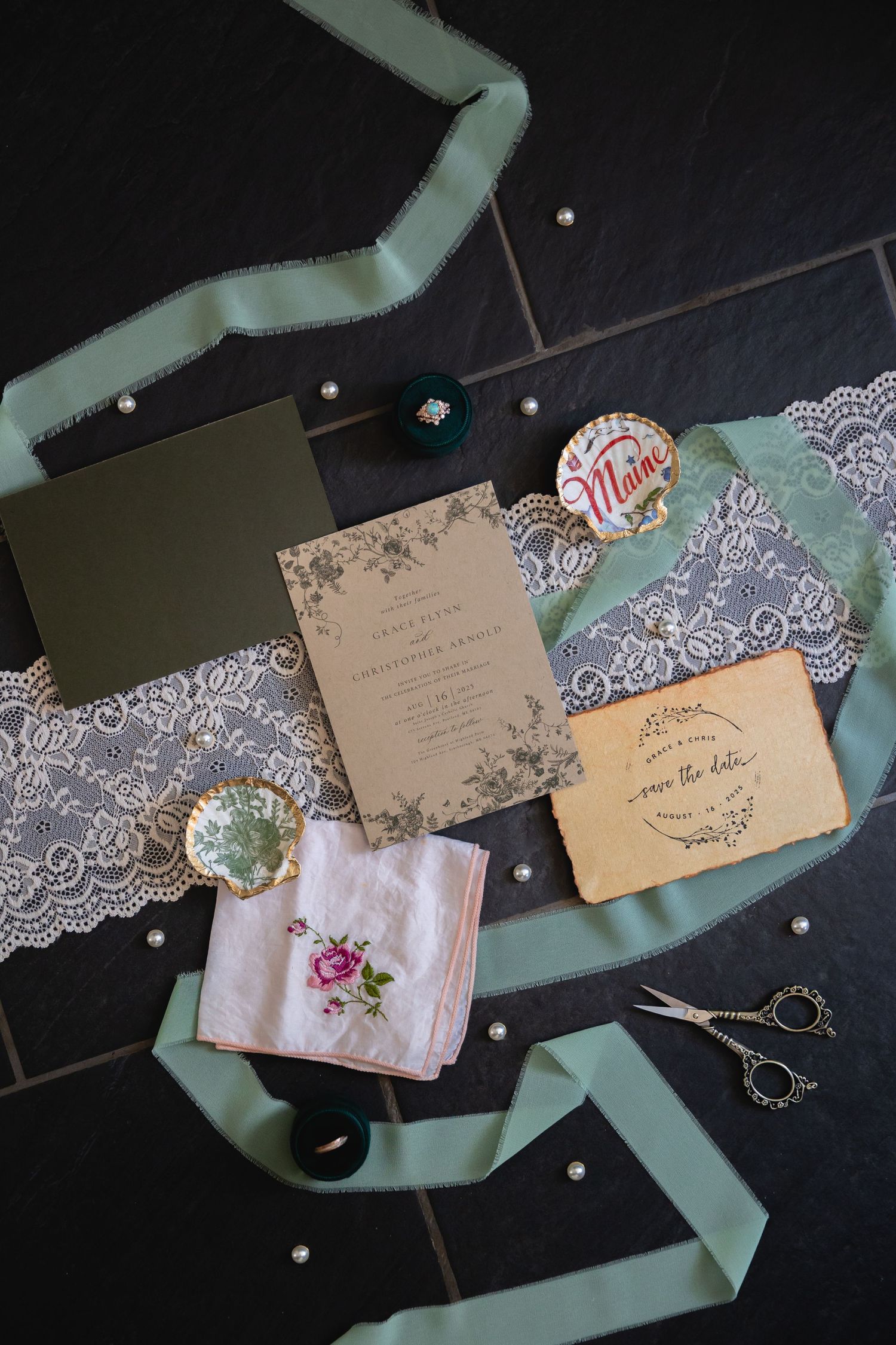 Wedding stationery flatlay with mint green ribbon, lace trim, vintage invitations and floral handkerchief on dark background.