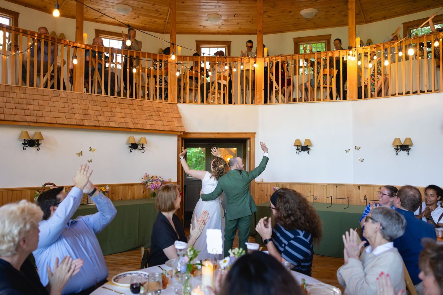 A group of people raise their hands in celebration during a gathering at a rustic restaurant with wooden balcony above.