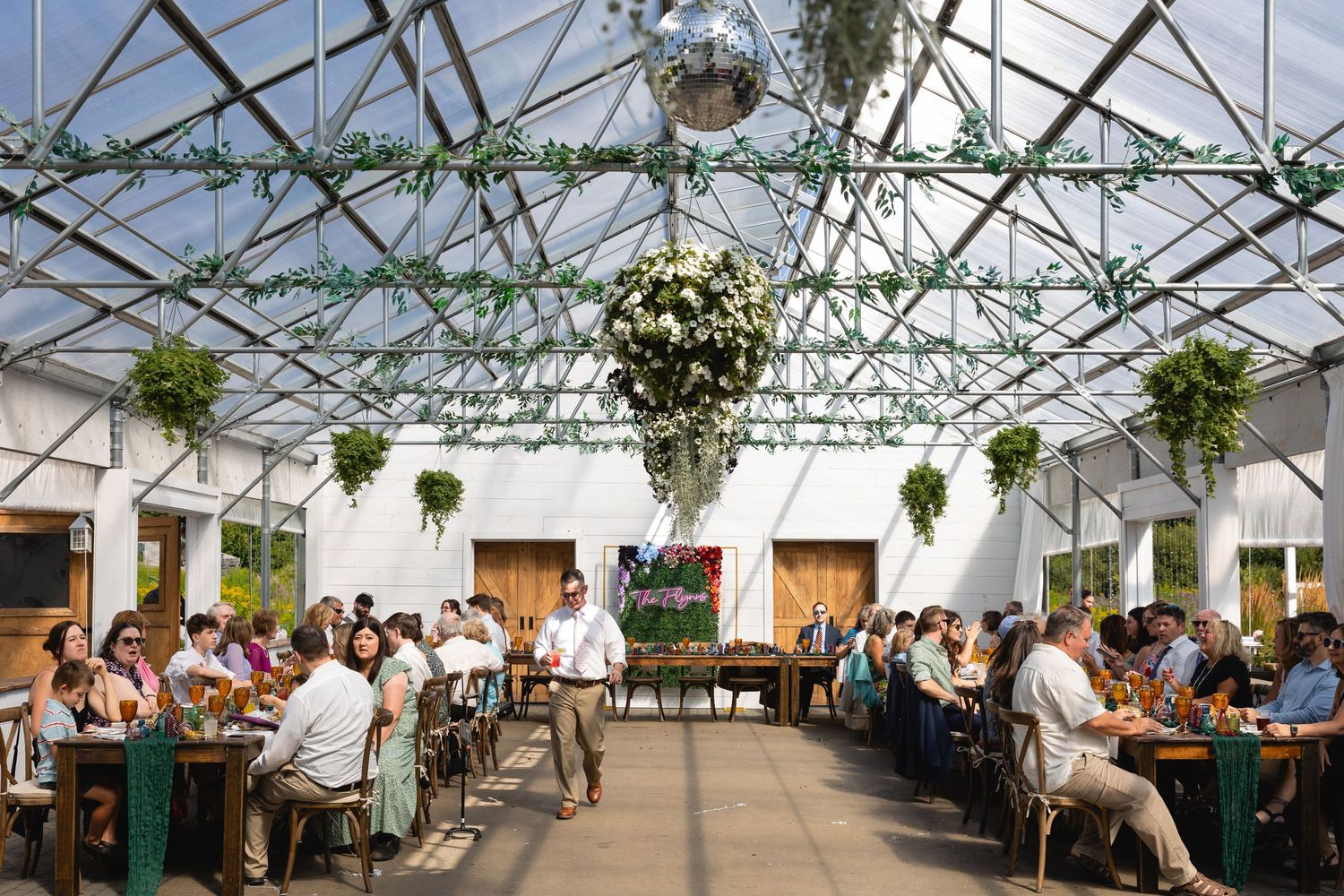 Wedding reception in a bright greenhouse venue with hanging disco ball and greenery decorations above seated guests.