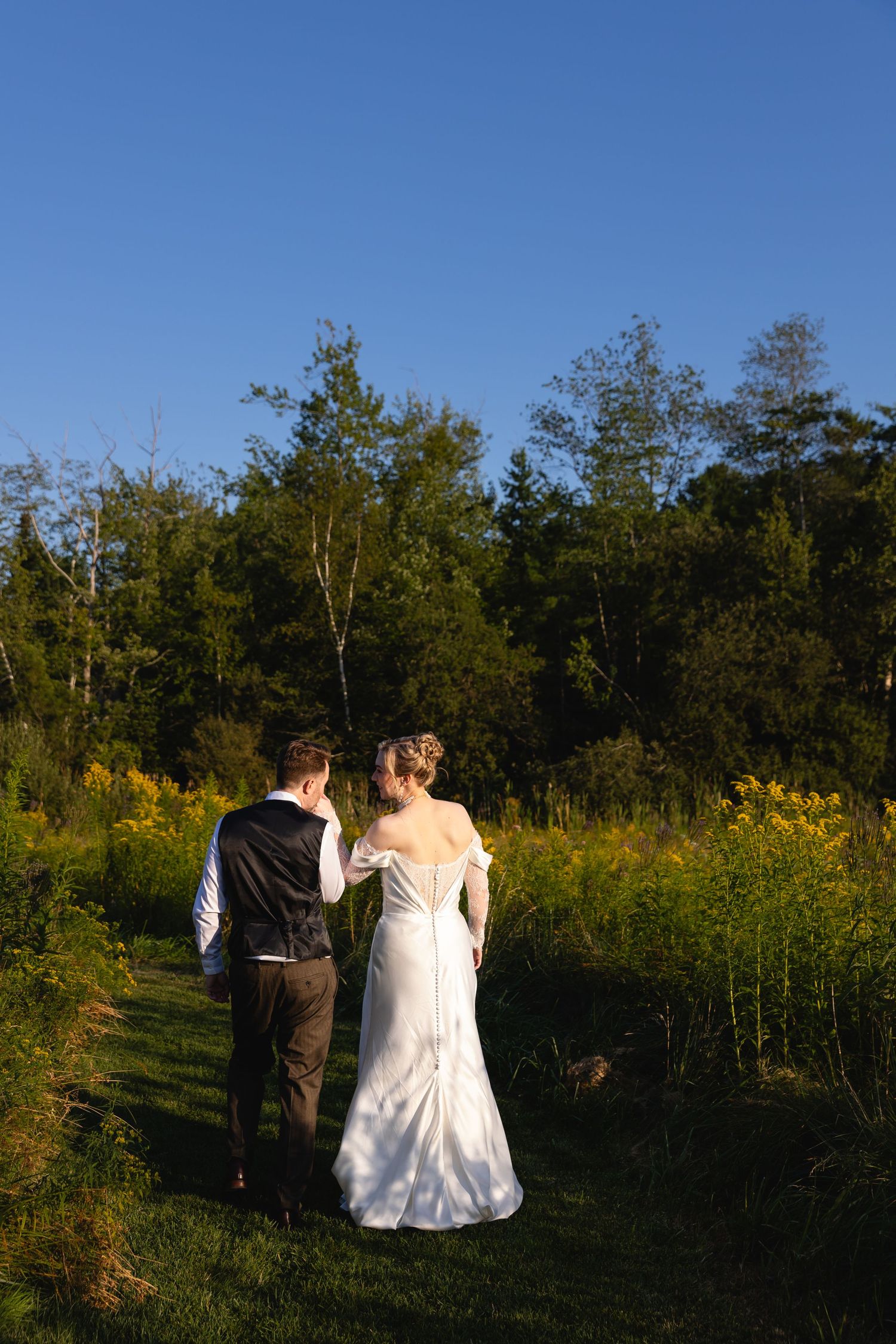 A couple in wedding attire sits together on a bench surrounded by autumn foliage and trees against a blue sky.