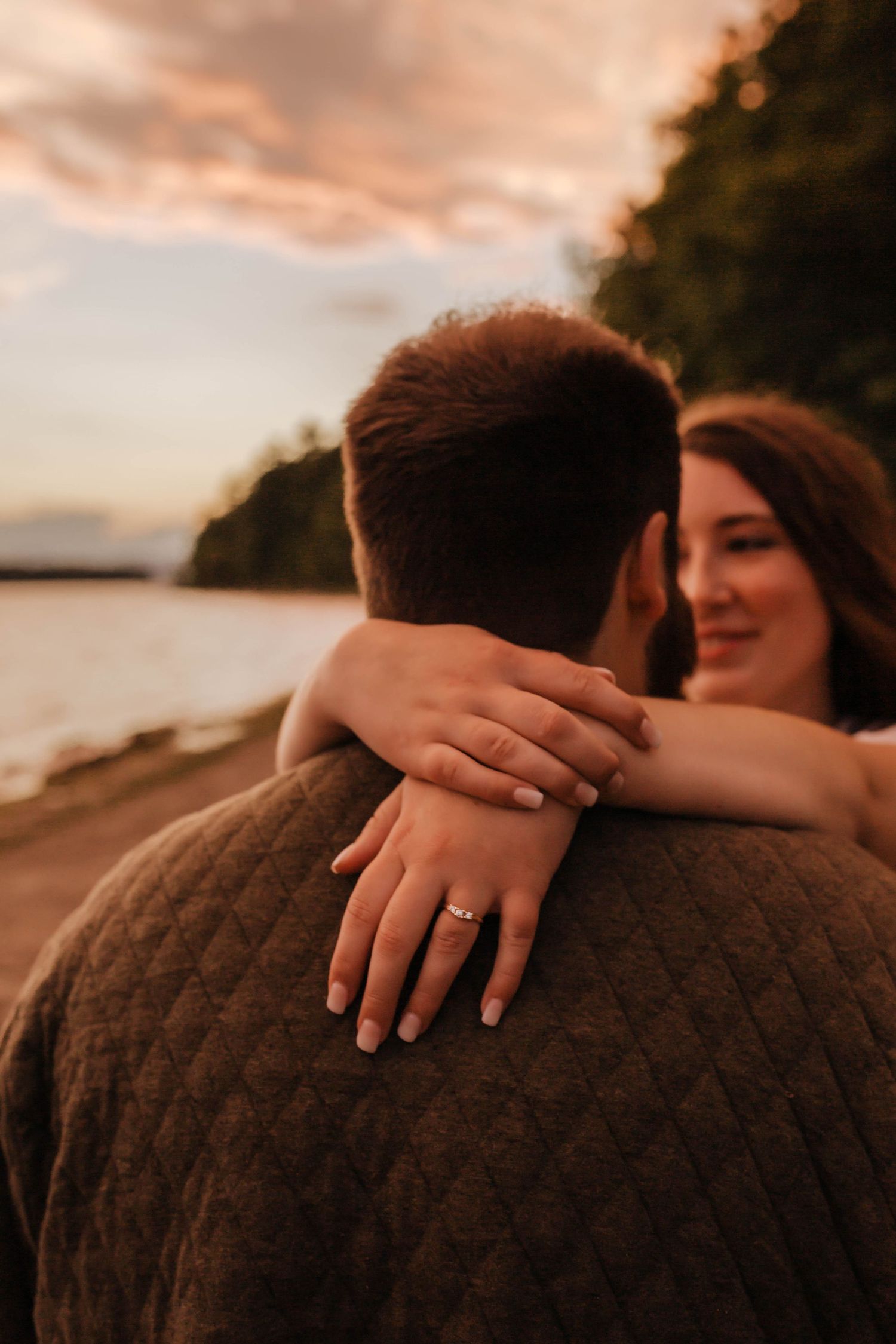 A romantic sunset embrace shows a close-up of hands with an engagement ring against a warm coastal backdrop.