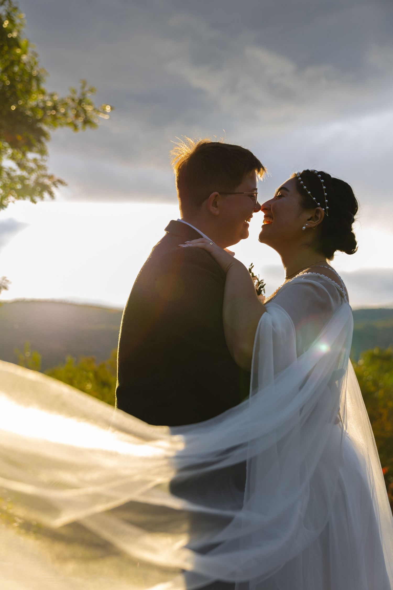 Wedding couple kisses as bride's veil flows in the wind with mountains and sunset in background.