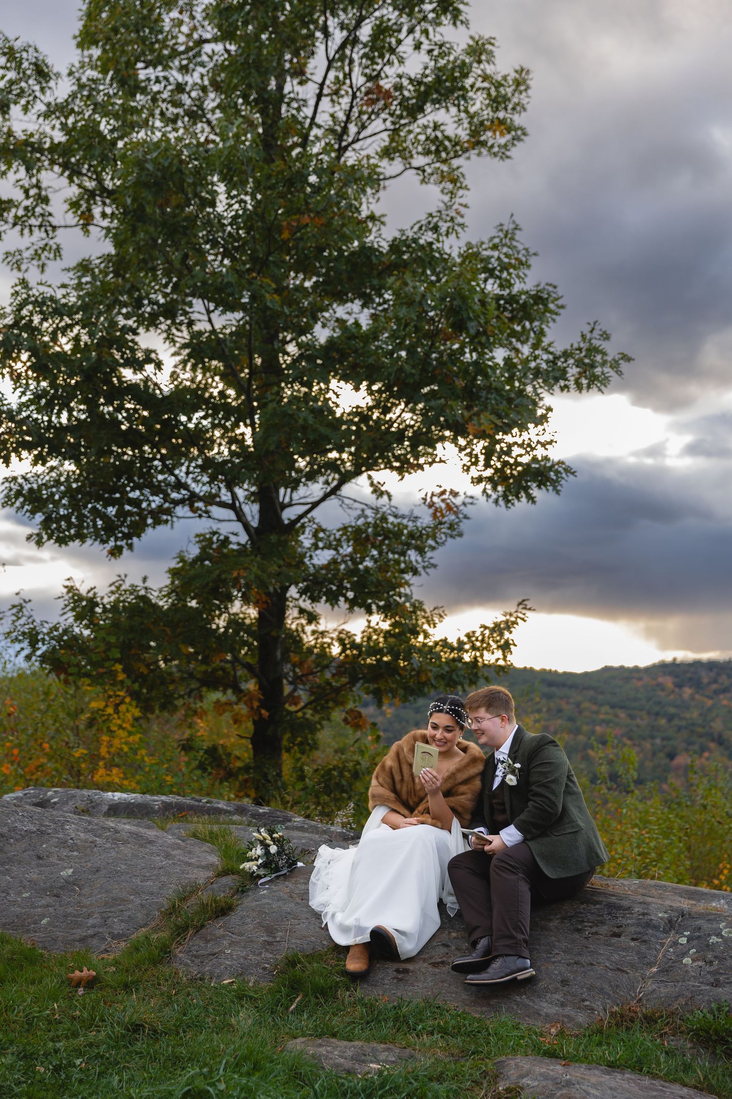 Couple in wedding attire sitting on rock formation with autumn mountain landscape in background.