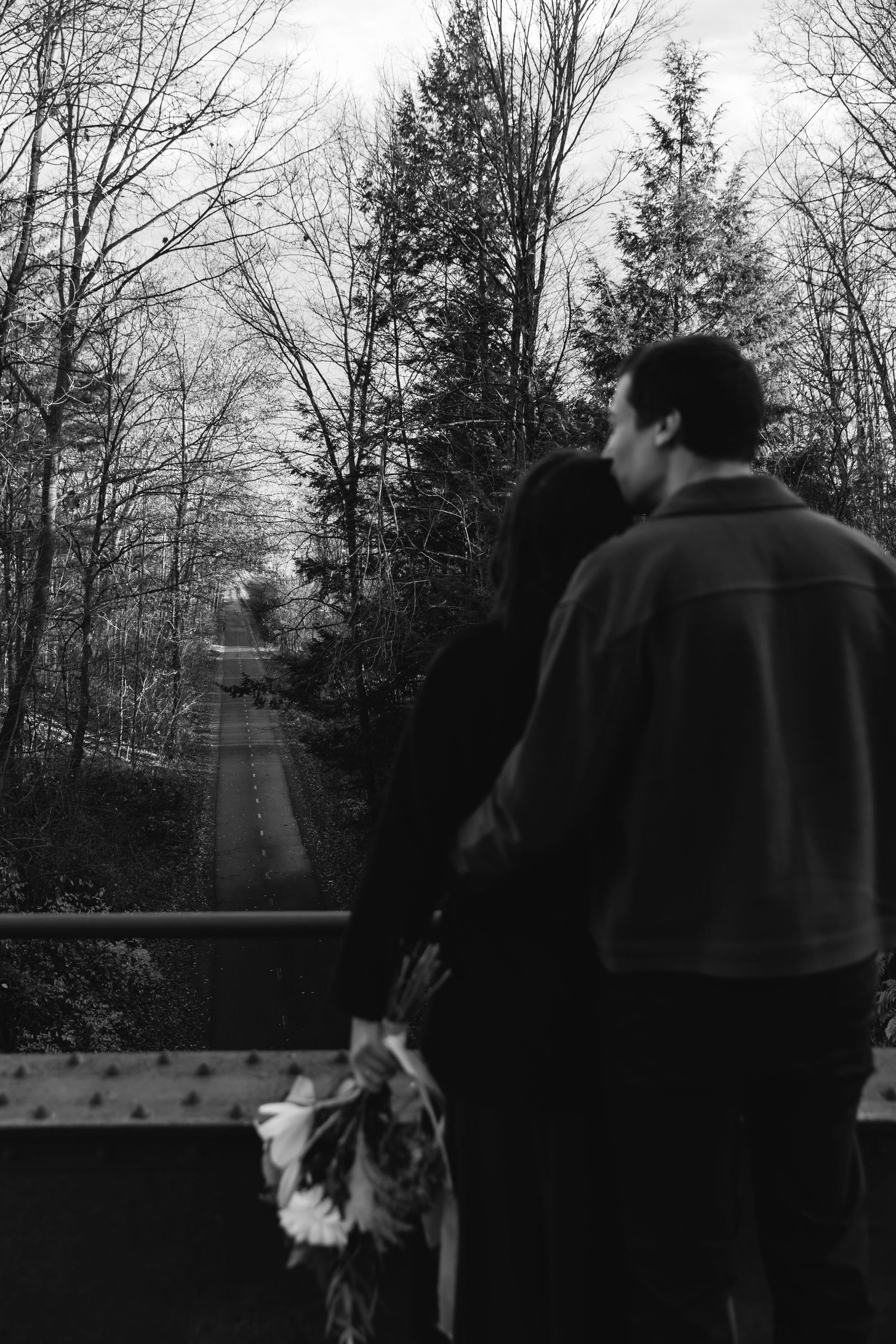 People standing on a bridge overlooking a winding trail through bare winter trees in a black and white scene.