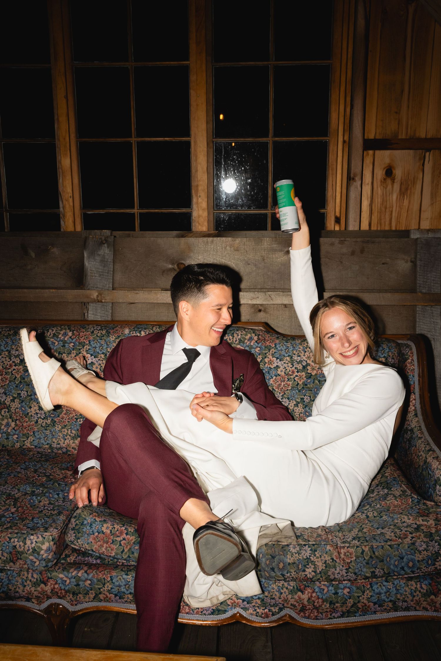 A playful moment on a floral couch showing two people in formal attire laughing and posing casually with drinks.