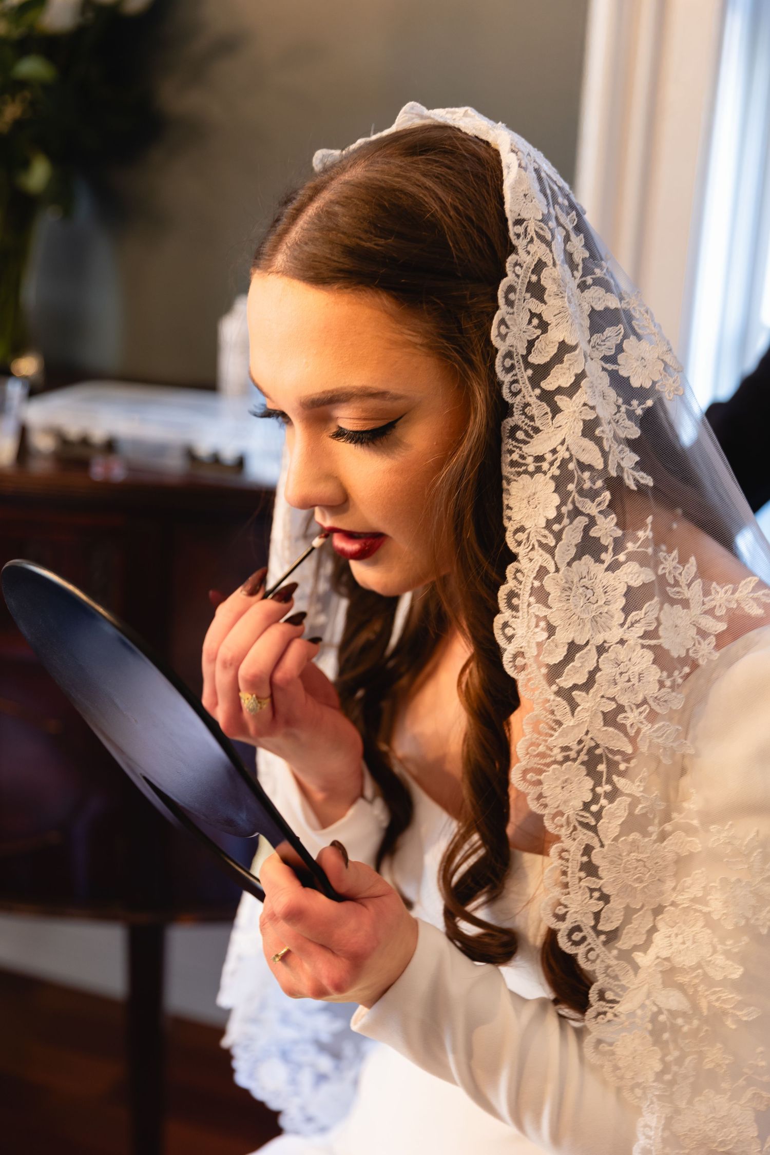 A bride in a white lace veil applies red lipstick while looking in a handheld mirror.