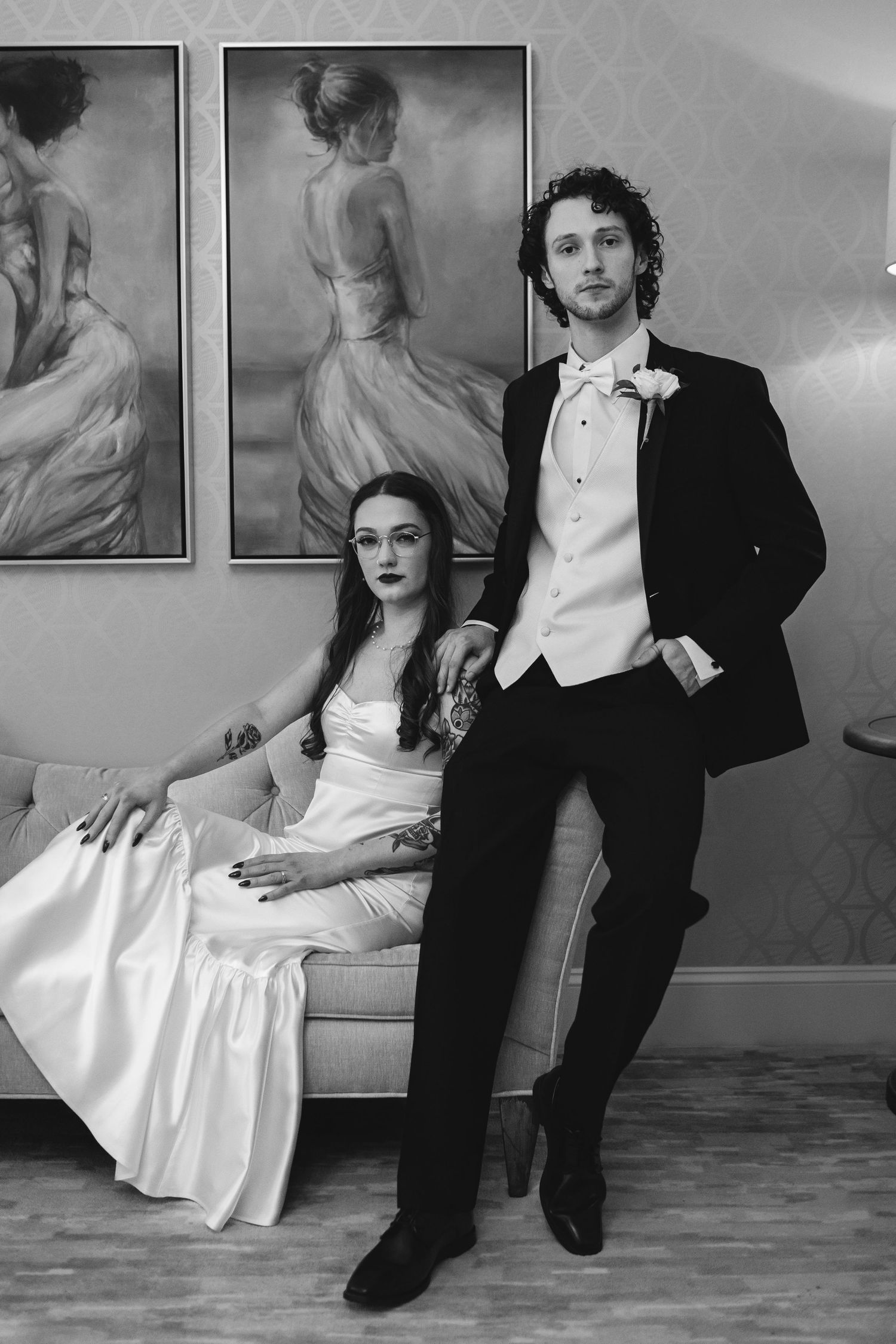 A formal black and white portrait of a couple in wedding attire posing against a wall with vintage artwork.