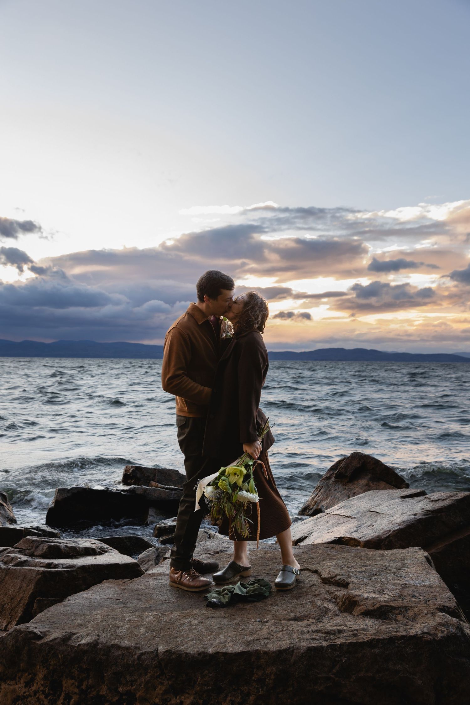 A couple shares an intimate moment on rocky coastal cliffs at sunset with dramatic waves crashing against the shore.