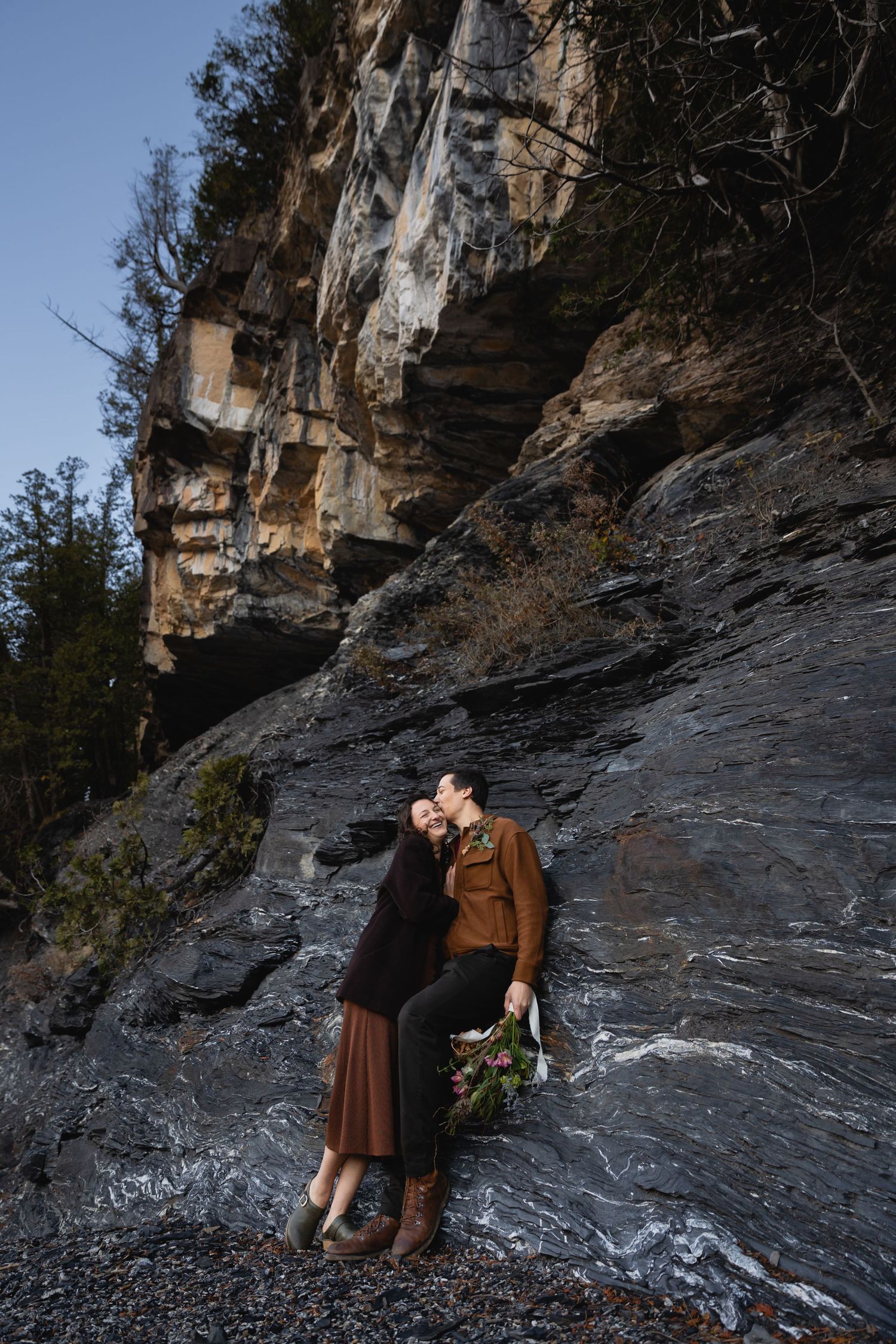 A couple shares a romantic moment against a dramatic rock cliff face while holding a rustic flower bouquet.