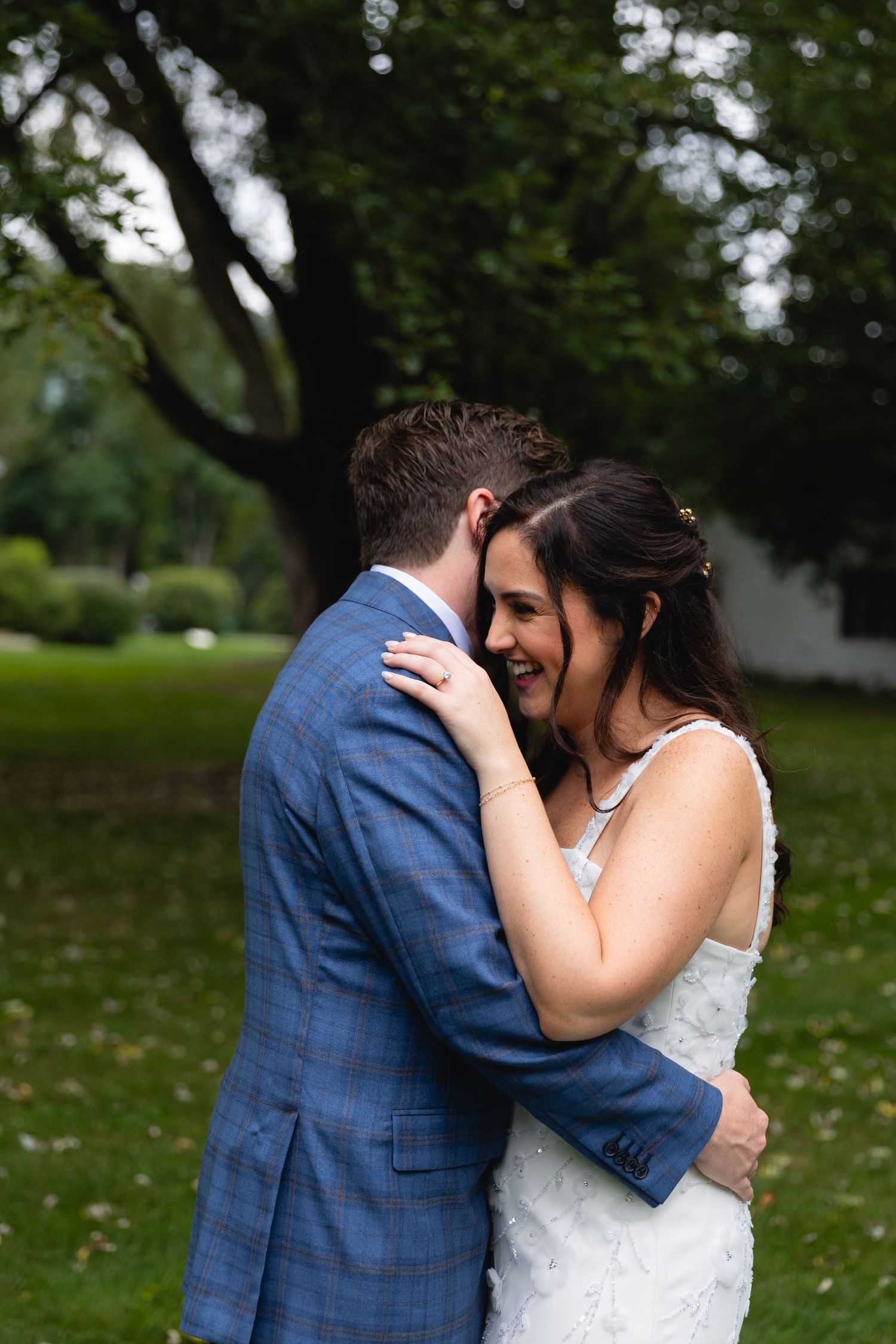 A bride in a white dress and groom in blue suit embrace lovingly outdoors under trees in a garden setting.