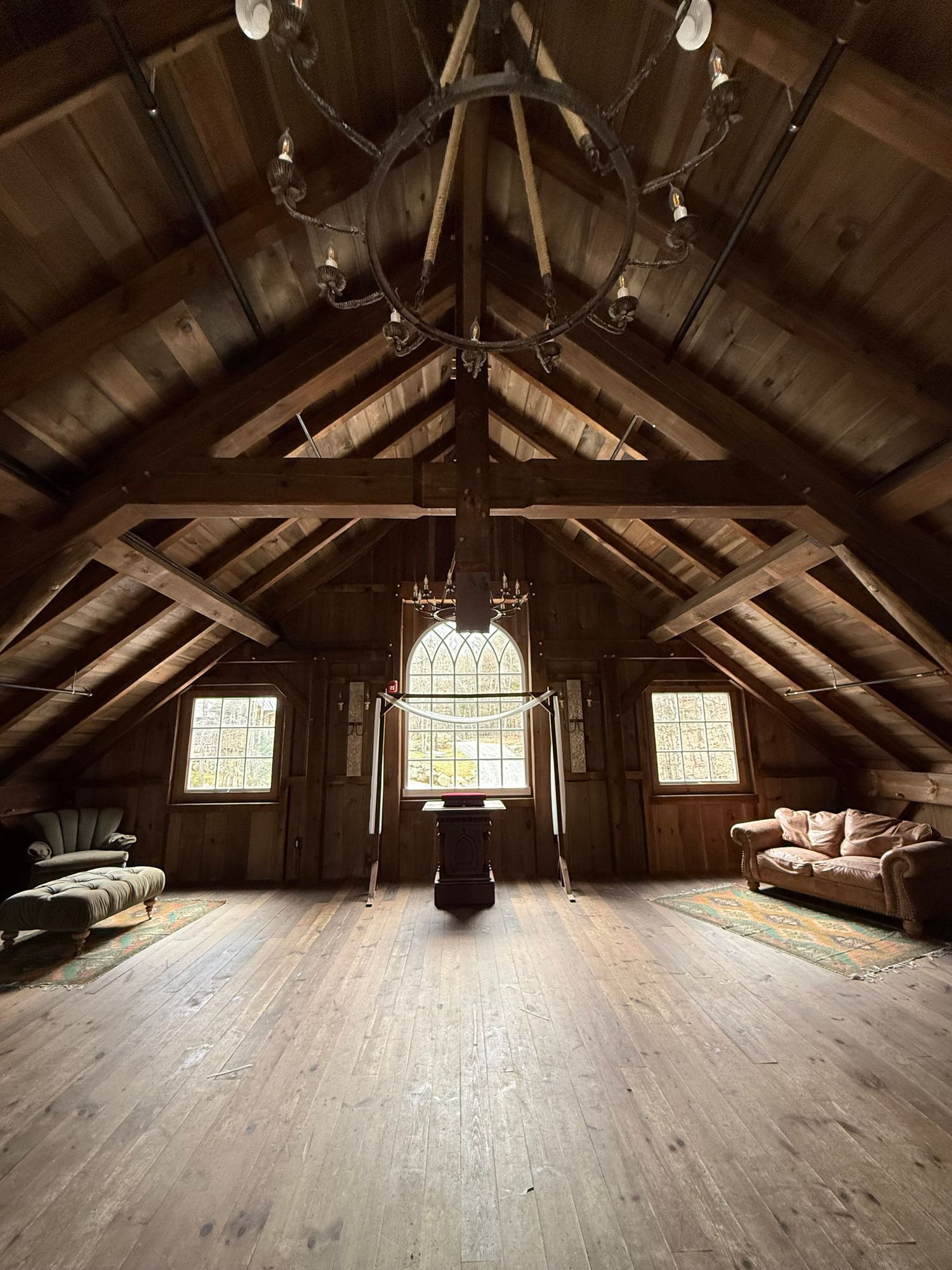A rustic attic room with exposed wooden beams, cathedral windows, hardwood floors and cushioned benches along the walls.