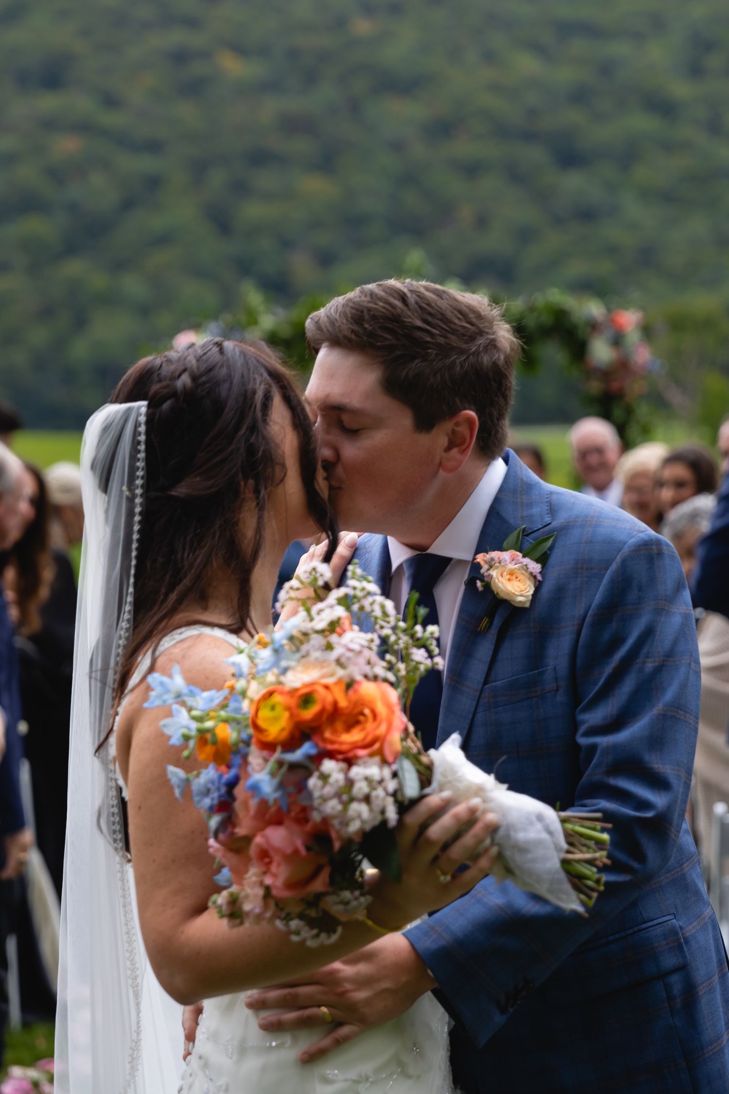 Newlyweds share a kiss during outdoor mountain ceremony while bride holds vibrant orange and blue wildflower bouquet.