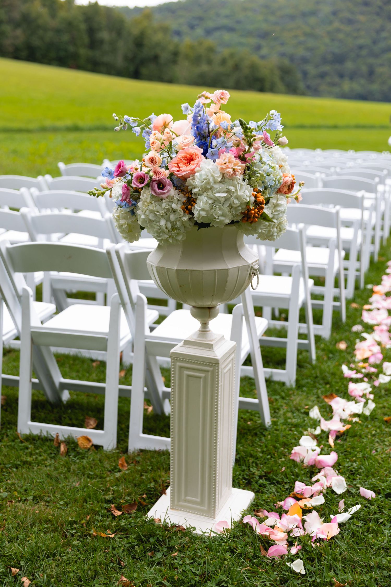 Elegant floral arrangement in white urn on pedestal with scattered rose petals and white chairs for outdoor wedding ceremony.