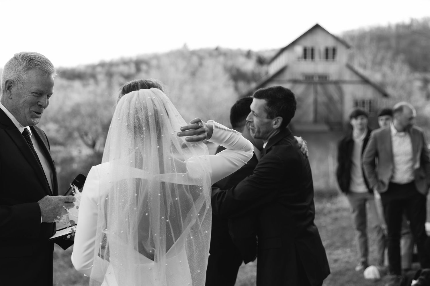 Black and white photo of an intimate outdoor wedding ceremony with bride in white veil and guests looking on.