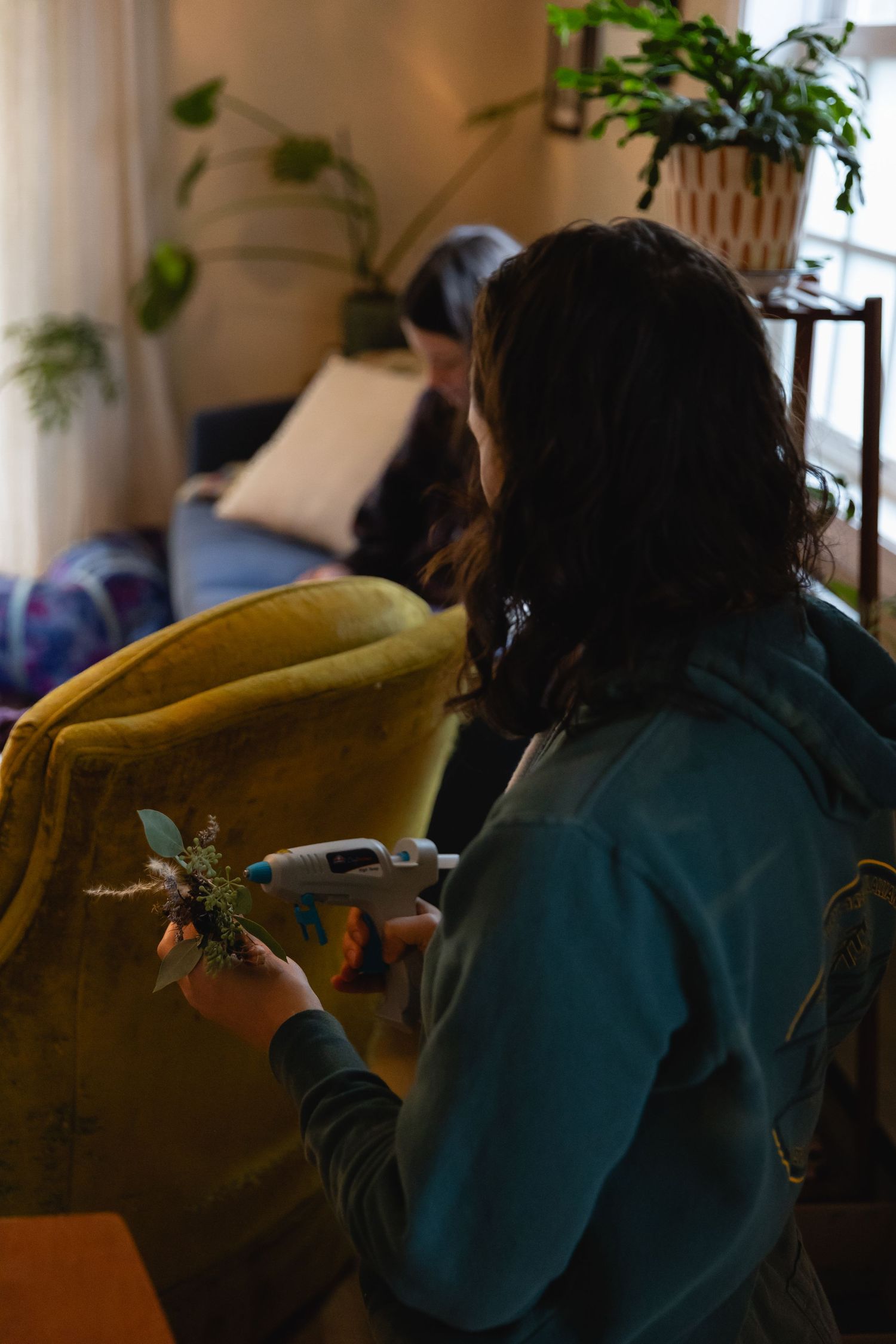 Person in teal sweater sits near houseplants while holding a device indoors.