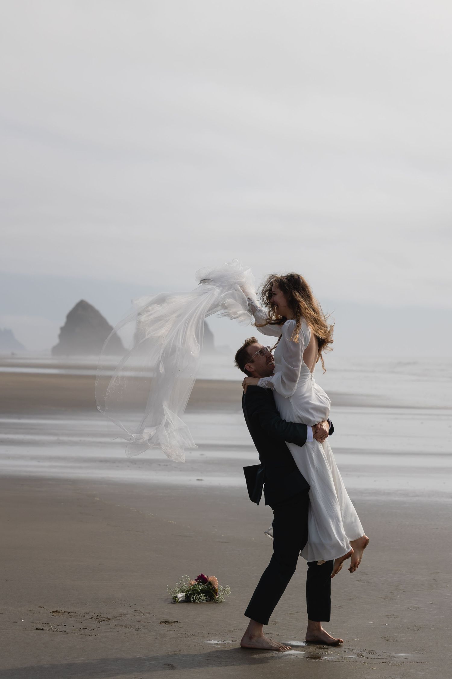 Groom lifts bride in white dress on a misty beach with Haystack Rock visible in the background.