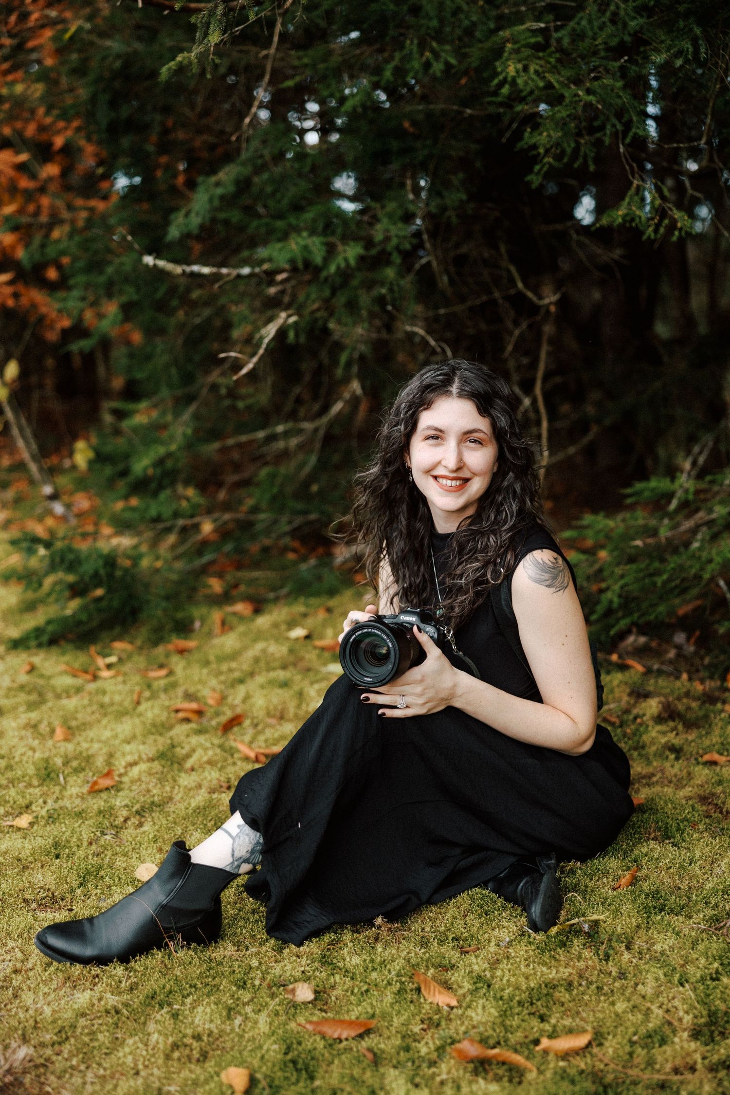 A smiling woman in a black dress sits on grass holding a camera, with autumn trees in the background.
