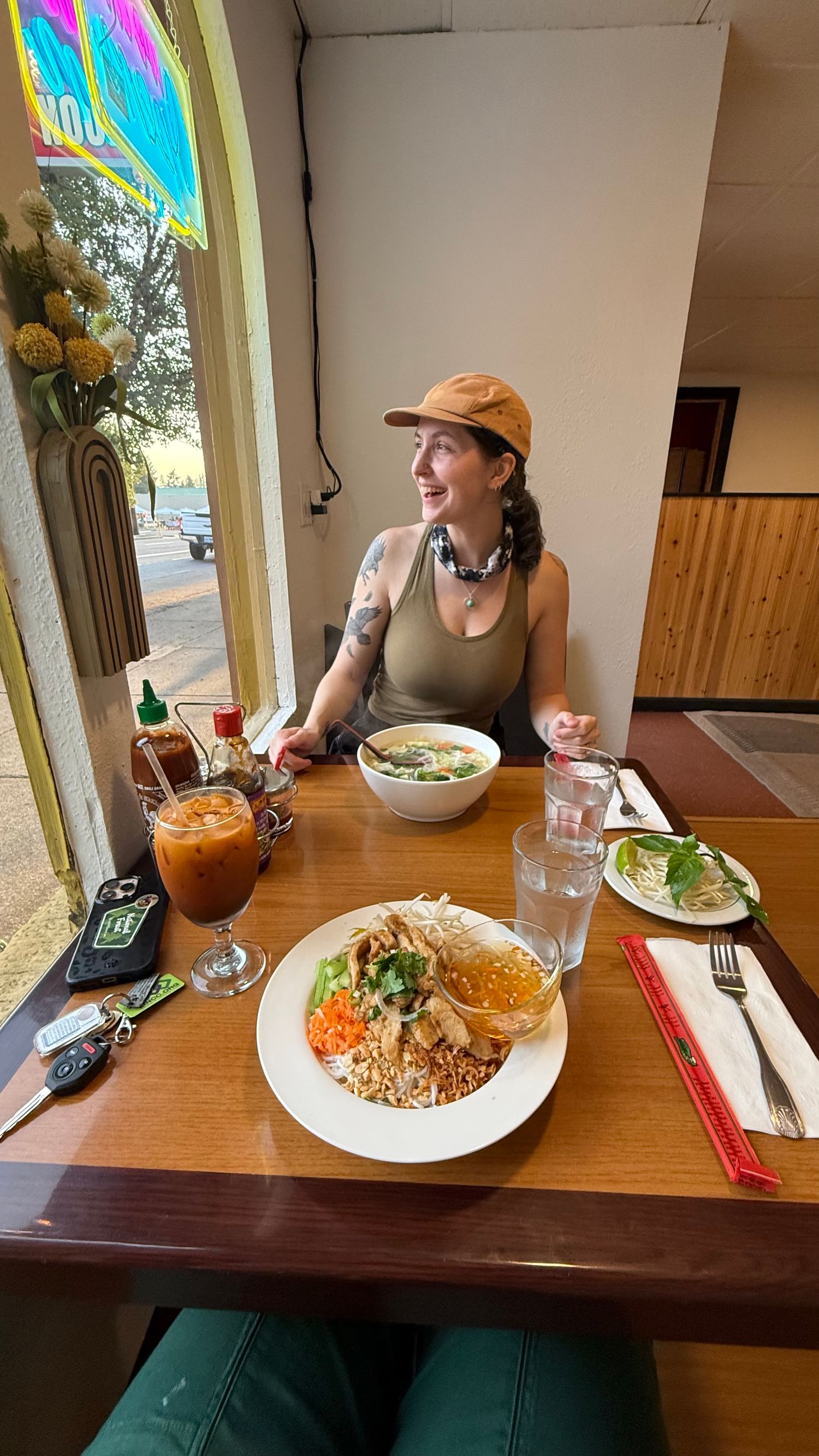 Woman in orange cap smiling at Vietnamese restaurant with pho and rice plate on wooden table.