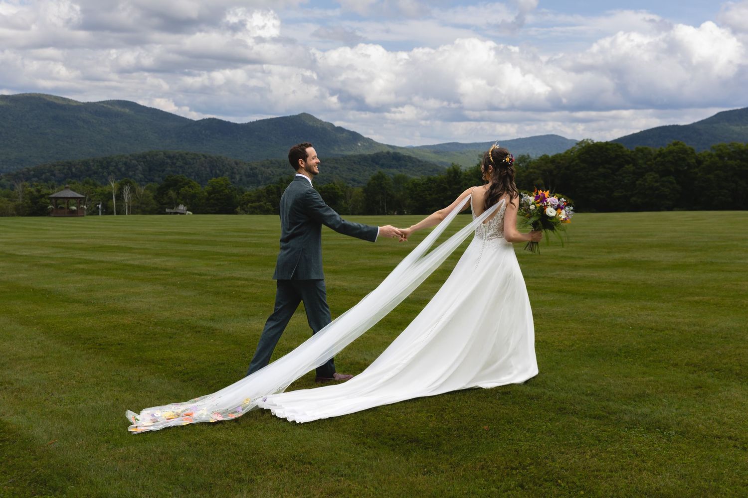 Wedding couple on green field with flowing veil and mountain landscape.