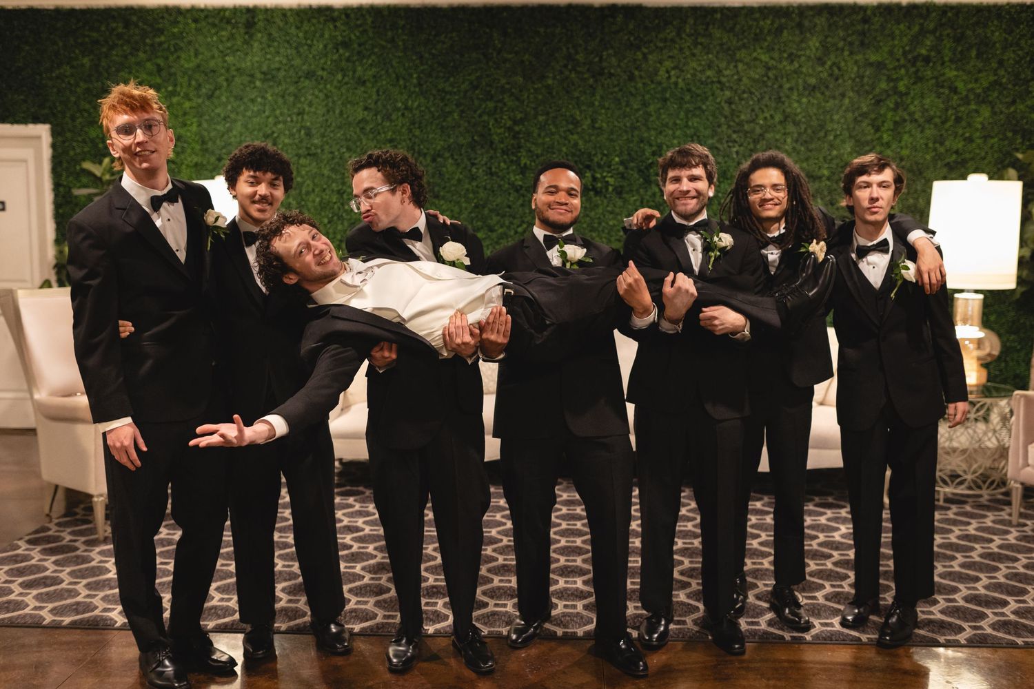 Groomsmen in black suits pose together for a fun group photo against a green hedge wall at a wedding celebration.