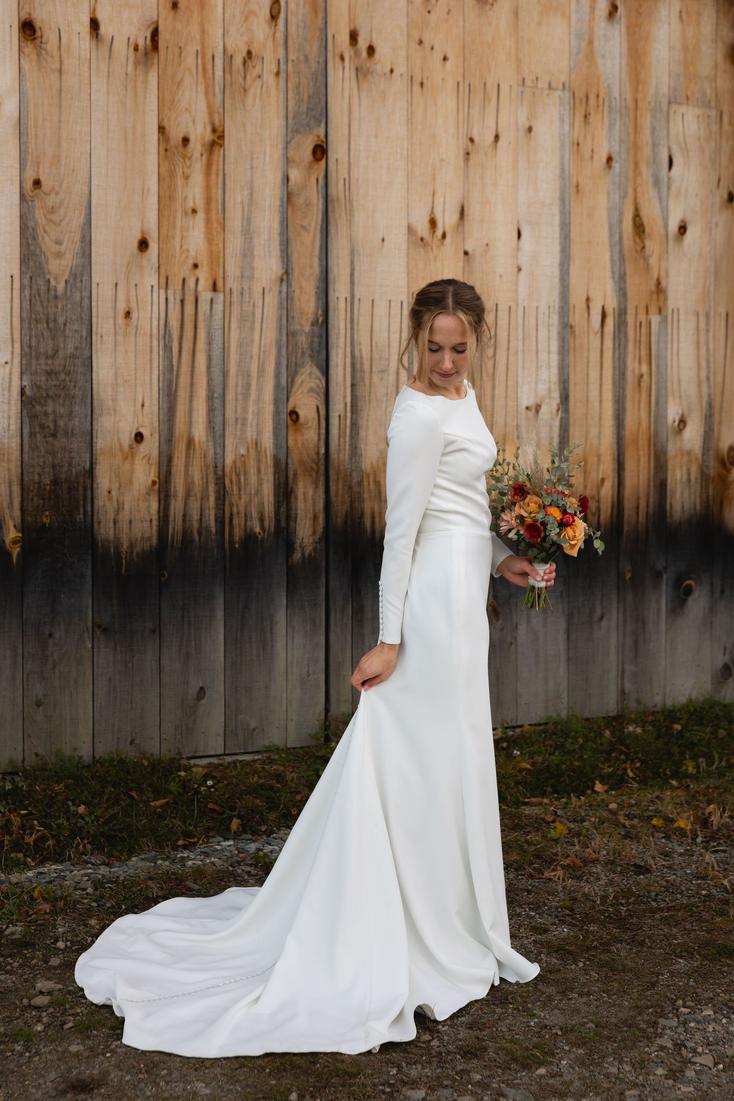 A white long-sleeved wedding dress with a train is showcased against a wooden fence backdrop, with colorful autumn bouquet.