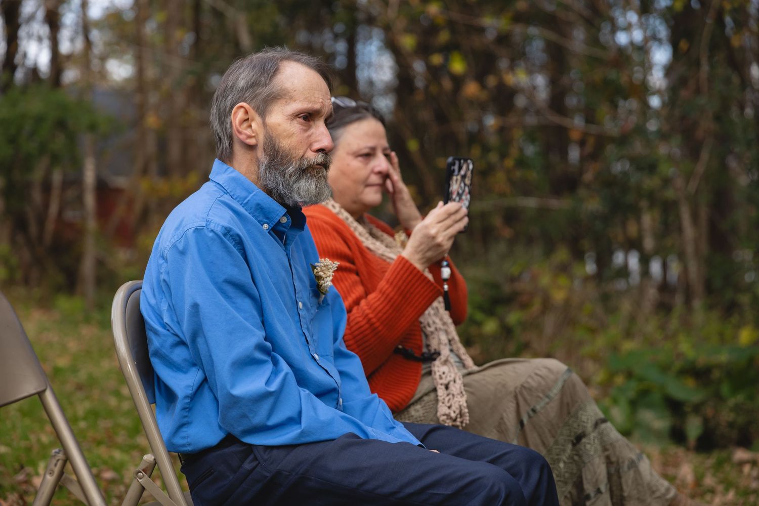 Wedding guests watching ceremony from folding chairs in wooded outdoor setting.