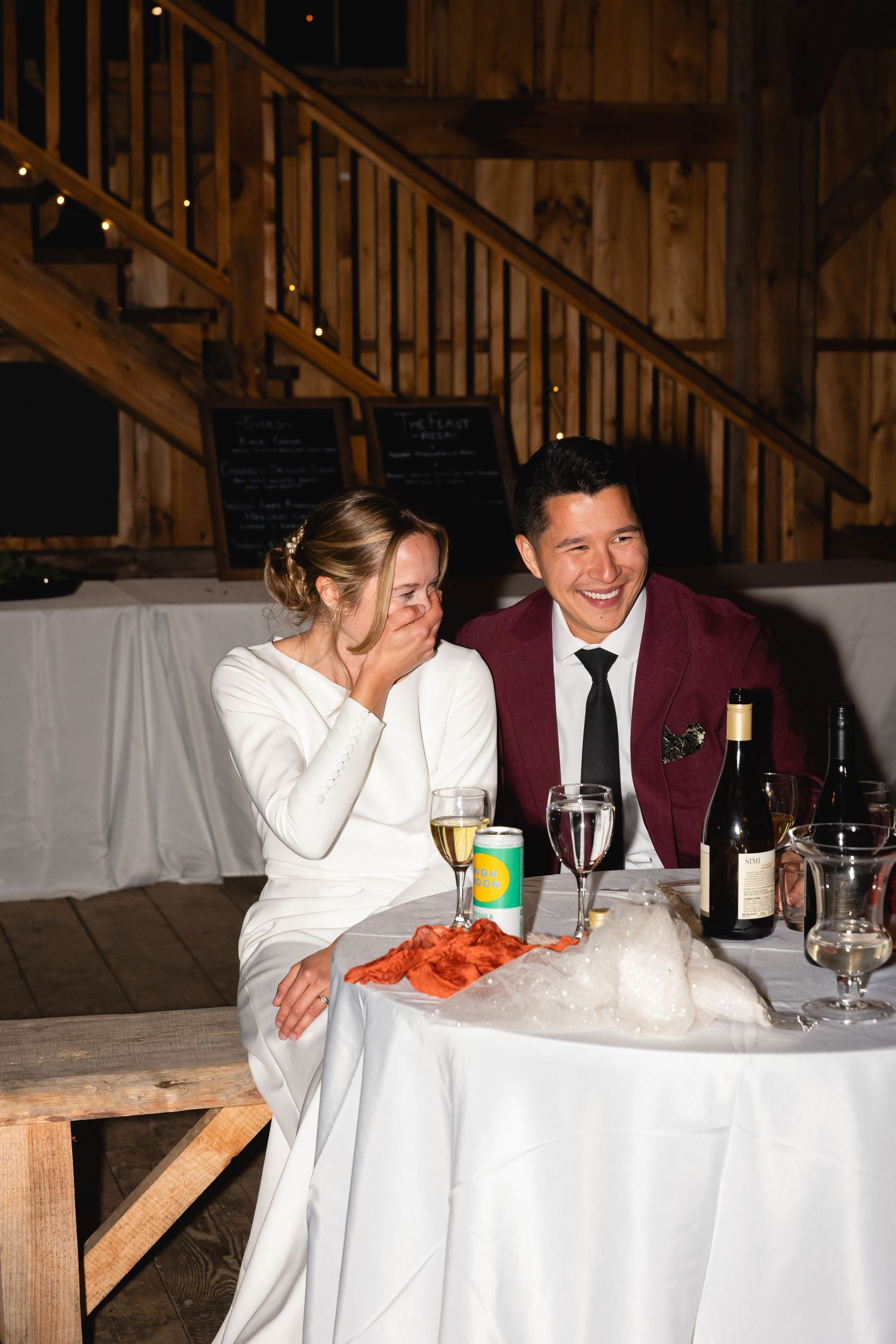 A couple shares a joyful moment at their rustic barn wedding reception with drinks and dinner on the table.