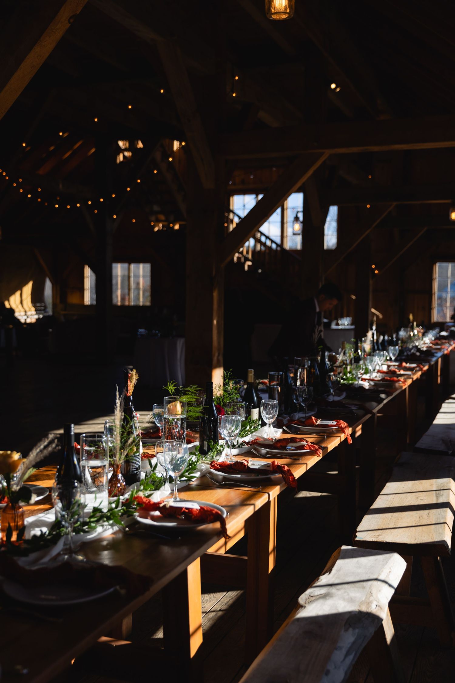 Long rustic wooden table elegantly set for dinner in a dark barn with string lights and moody ambiance.