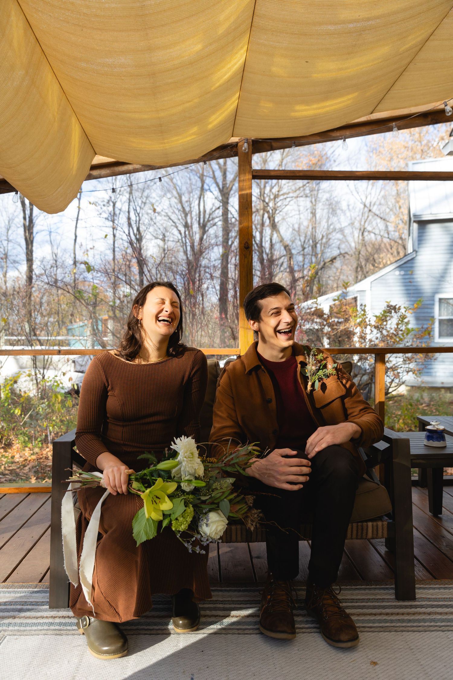 Two people in brown clothing share a laugh while sitting on a wooden deck with white flowers under a yellow canopy.