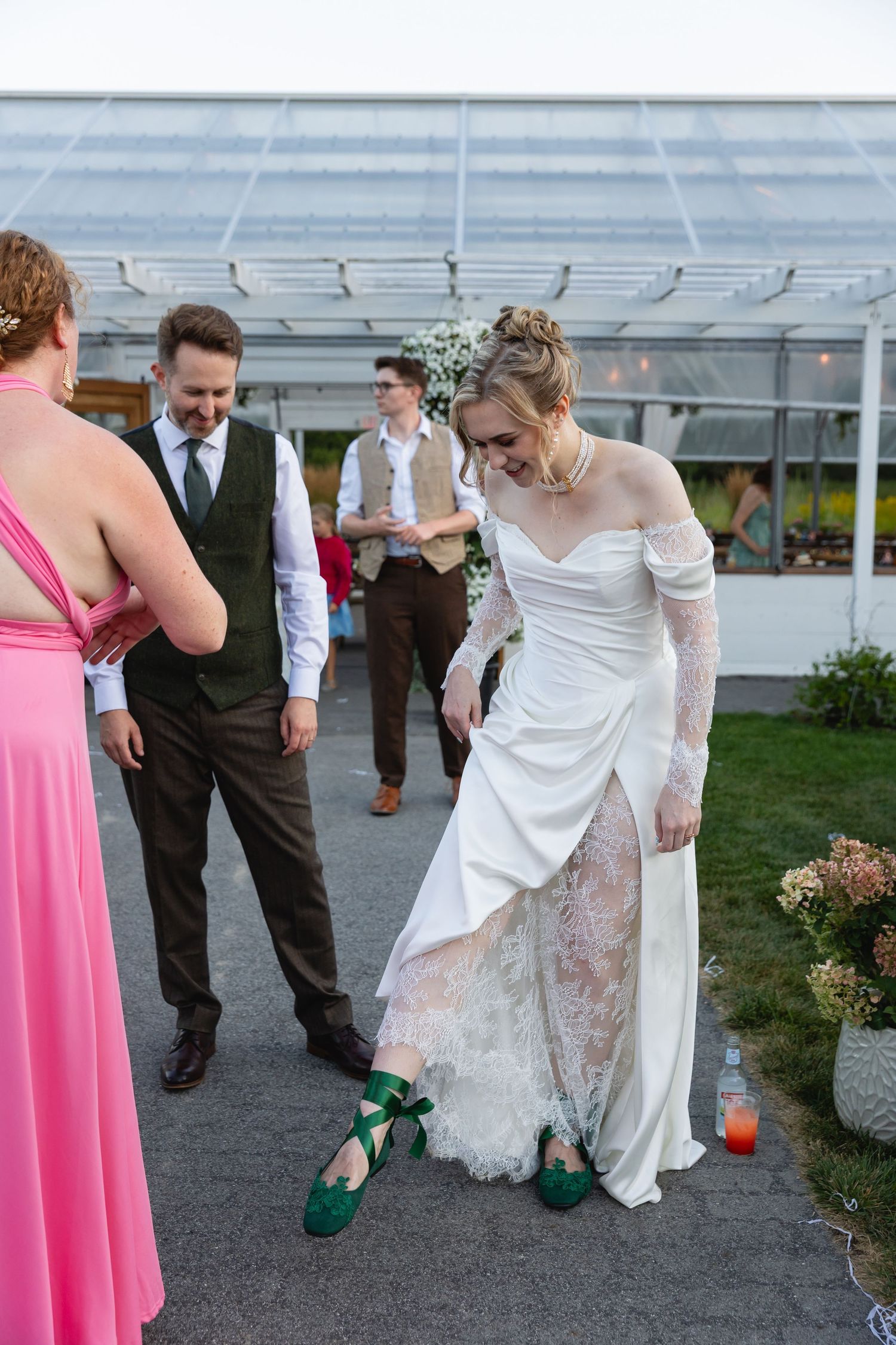 Wedding party gathered outside a greenhouse as bride adjusts her elegant white dress and green shoes.