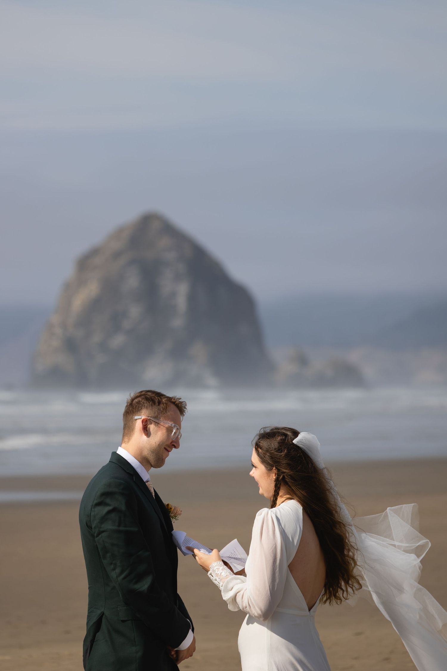 A couple stands on a misty beach with dramatic rock formations in the background during a wedding photo session.