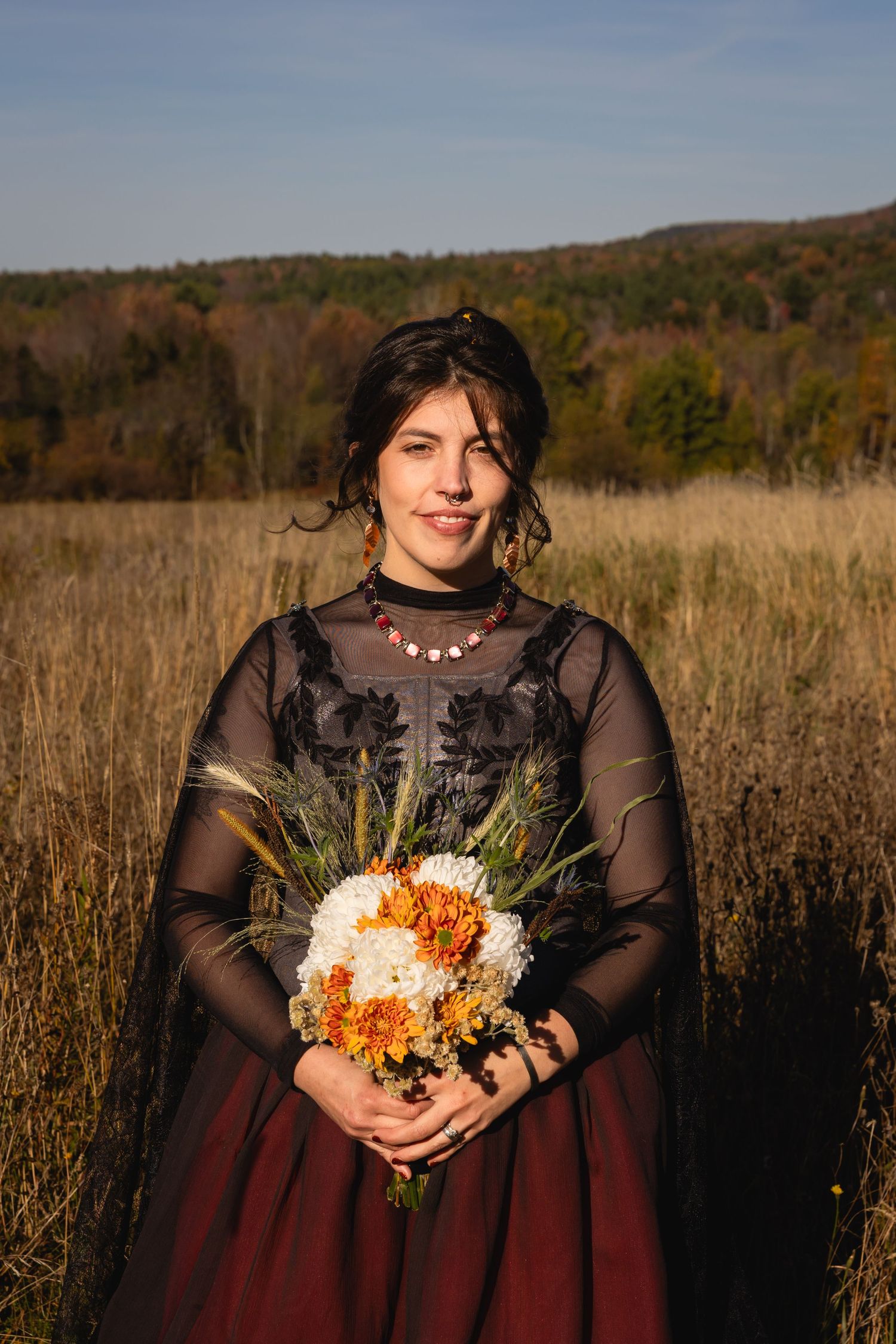 A figure in a burgundy and black dress holds an autumn-colored bouquet while standing in a golden field at sunset.