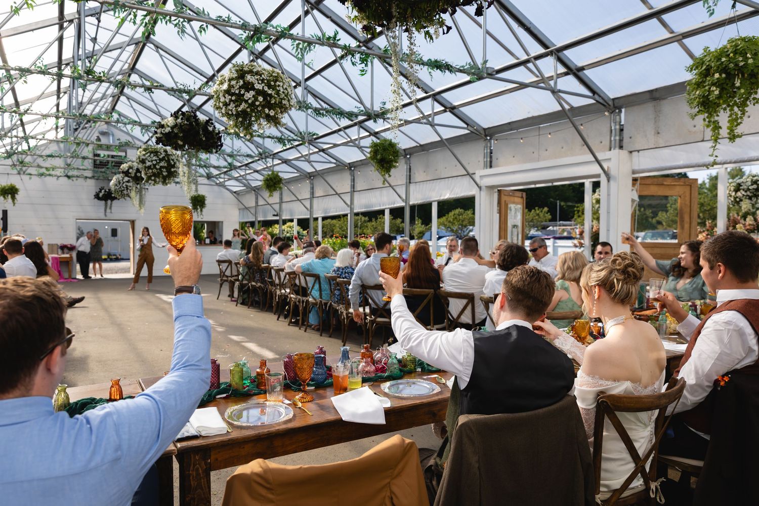Wedding celebration in a greenhouse venue with guests seated at long tables under glass ceiling.