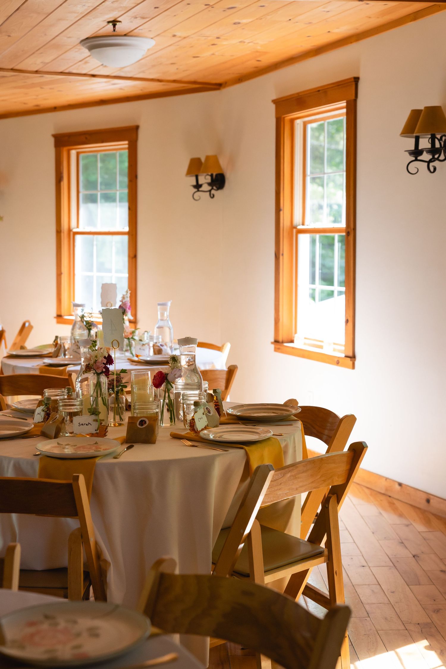 A rustic dining room with wooden beams, natural light, and round tables set for a special event.