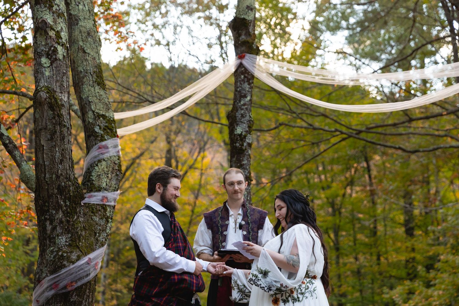 A wedding ceremony takes place under draped fabric in a wooded setting during autumn.