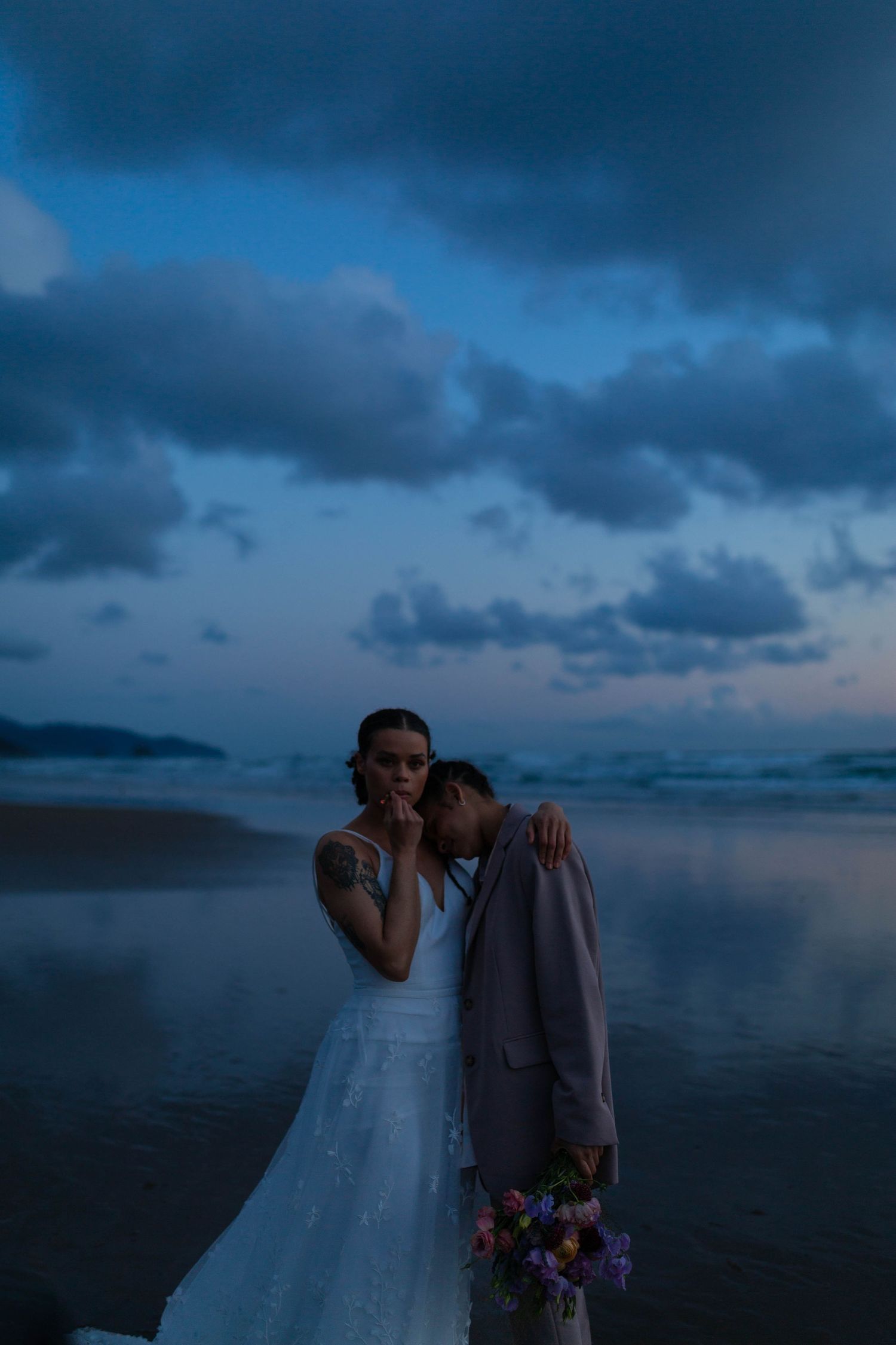 Two people embrace on a moody beach at sunset with dark clouds and ocean waves in the background.