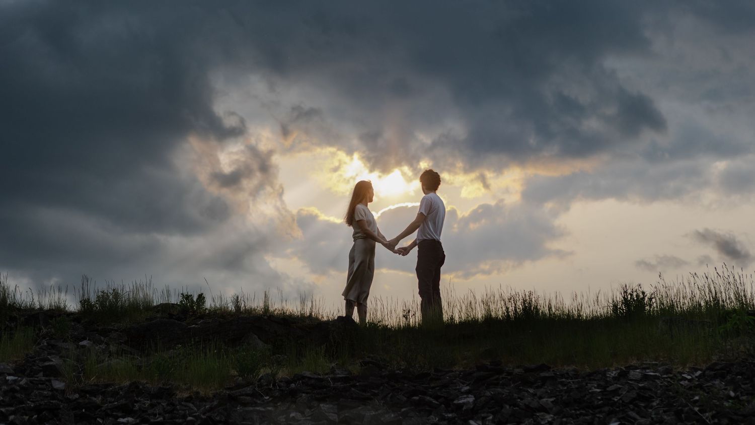 Couple holding hands in silhouette against a dramatic sunset sky with glowing clouds on a hilltop.