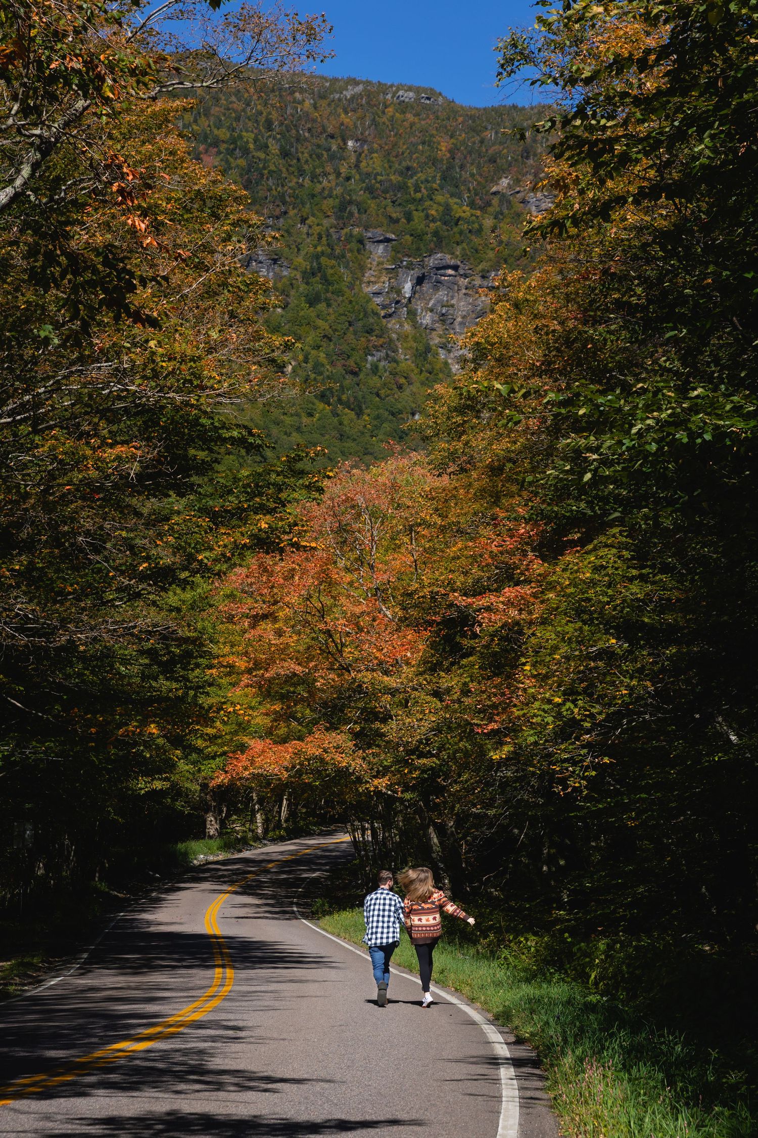 Couple walking hand in hand on a scenic autumn road surrounded by colorful fall foliage and mountain views.