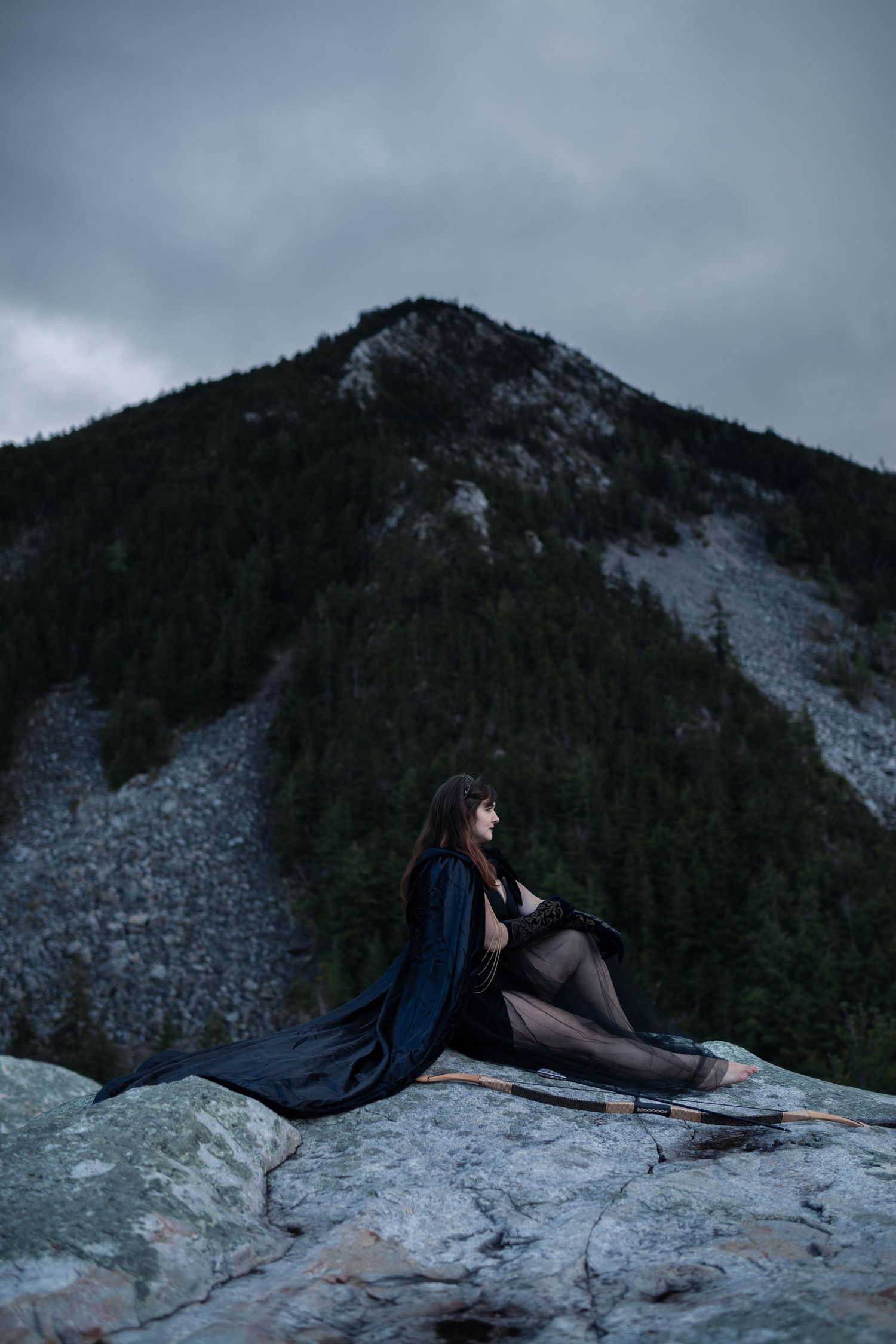 A woman in a dark flowing cape sits on rocky terrain with a dramatic mountain and forest backdrop.