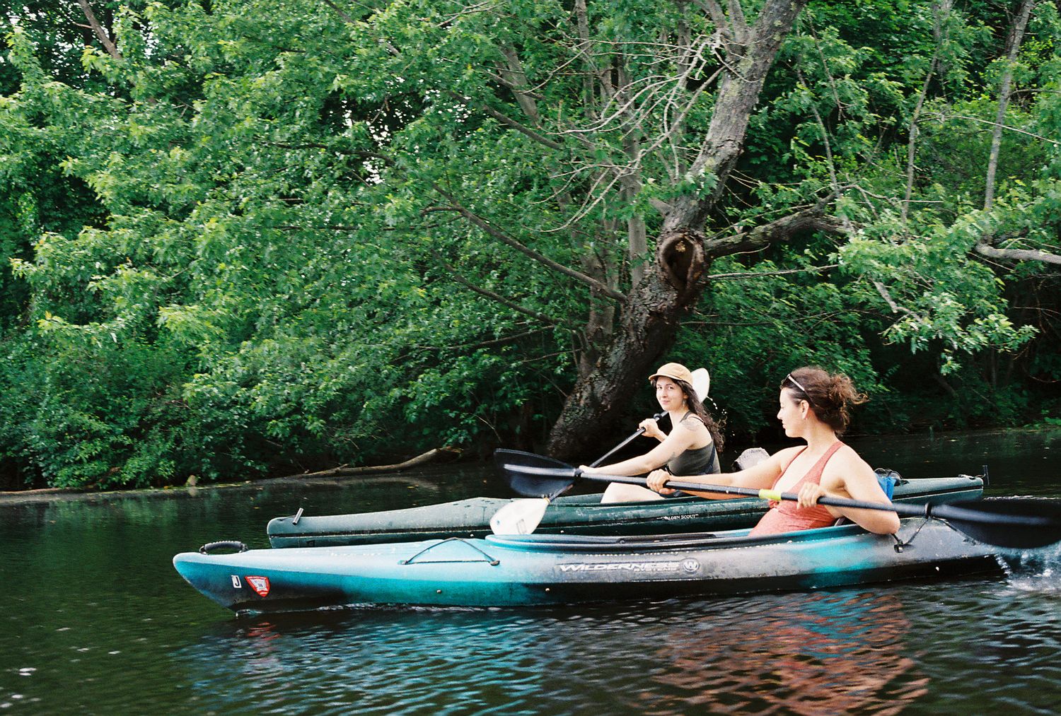 Two women paddle kayaks on a calm river surrounded by lush green trees in summer.