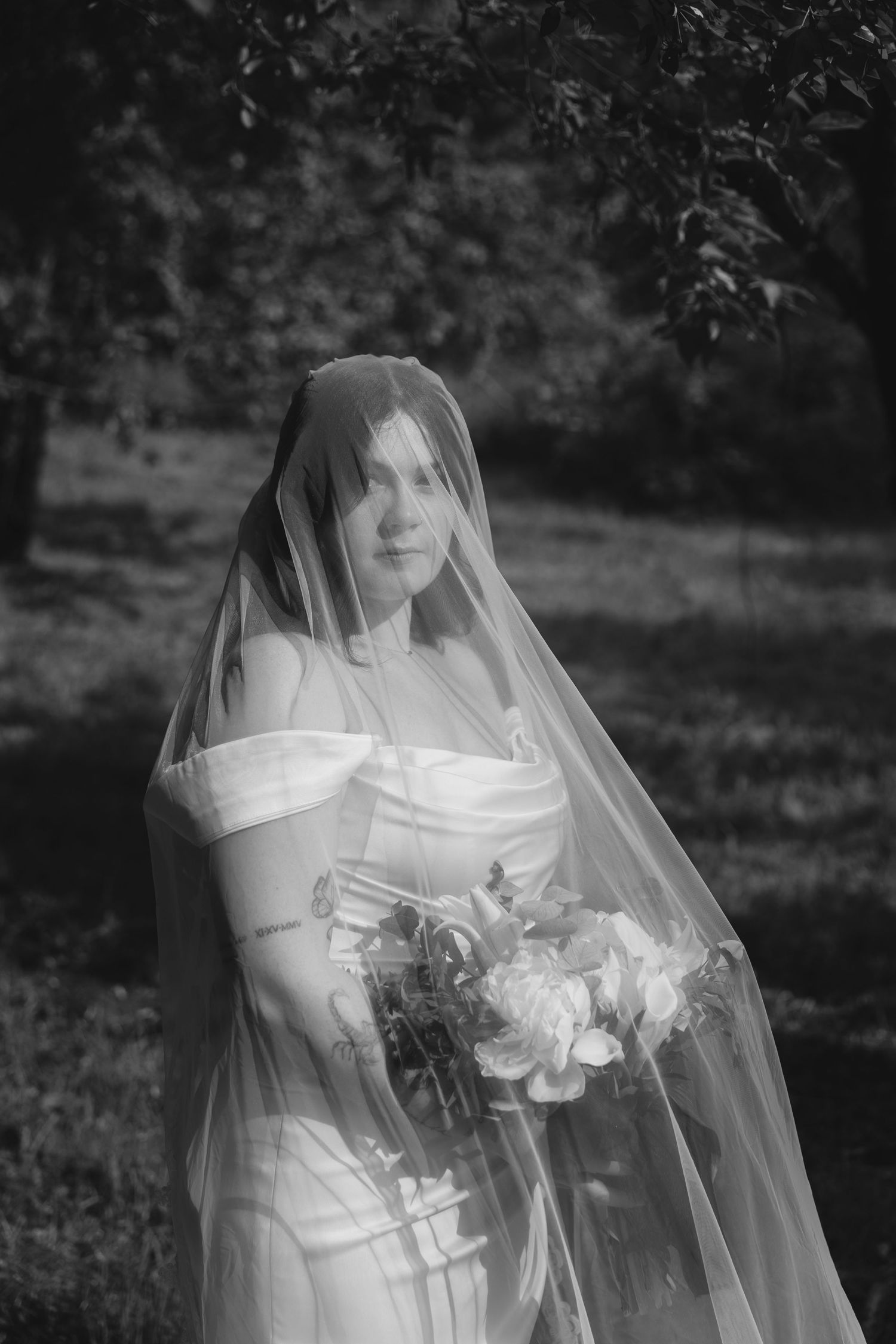 Elegant bride in white off-shoulder gown and veil holds bouquet outdoors in black and white wedding portrait.