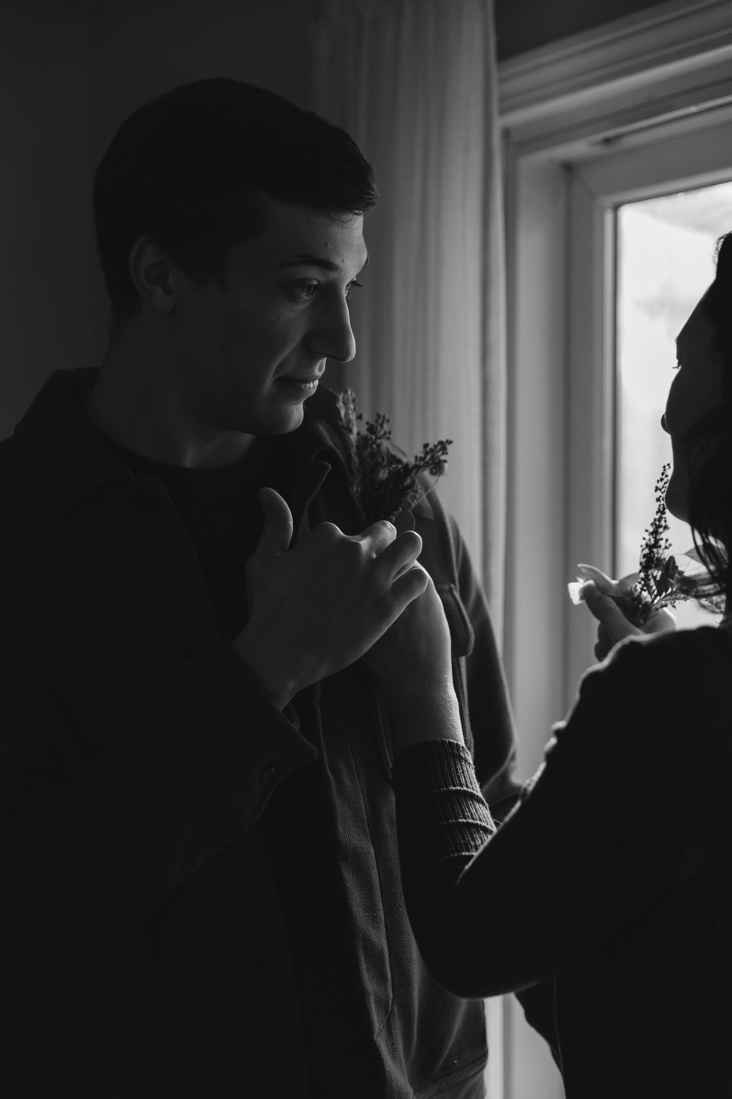 A black and white portrait of two people engaged in conversation near a window, creating a moody silhouette effect.