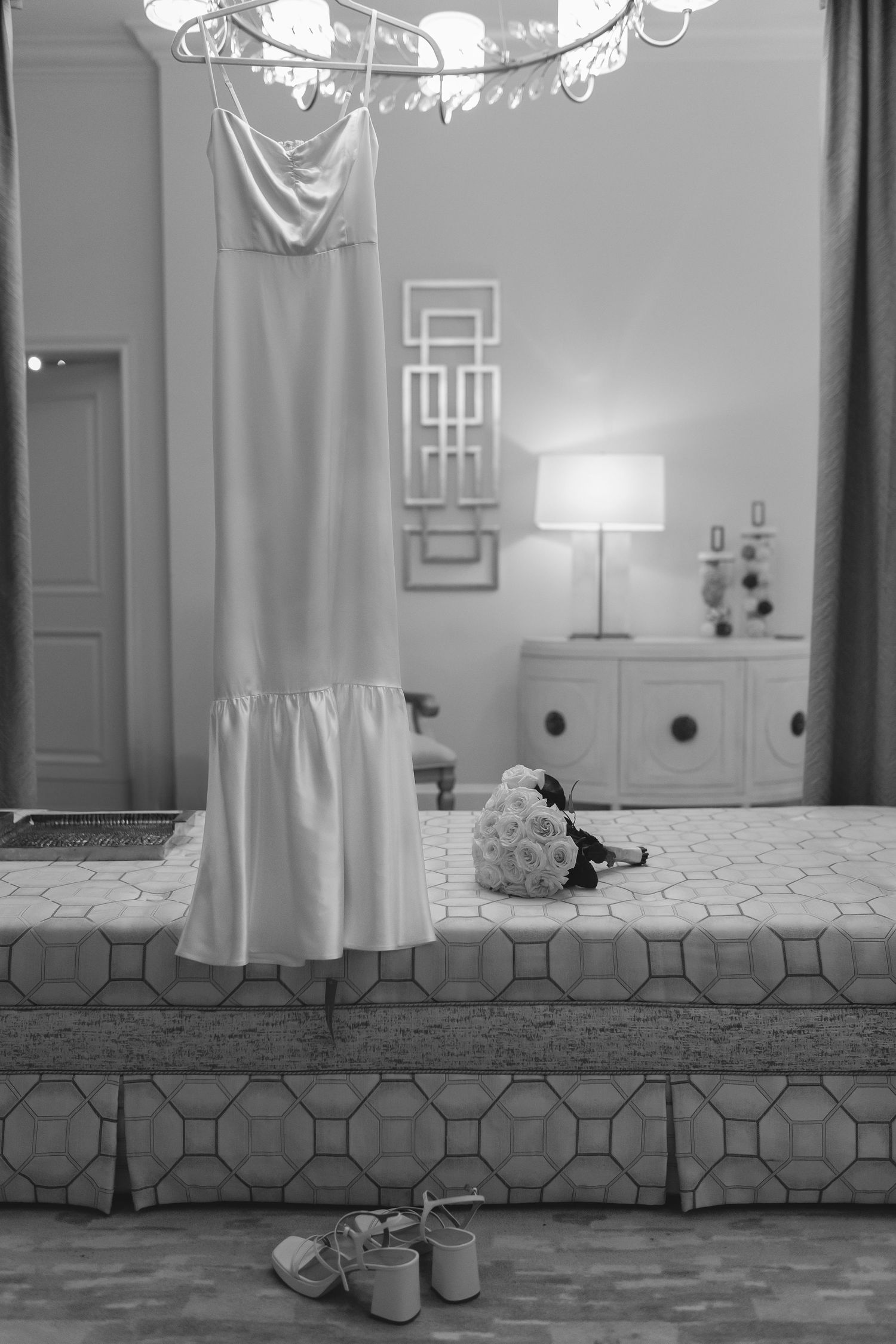 A white wedding dress hangs elegantly beside a table lamp, with bridal shoes placed on the wooden floor below.