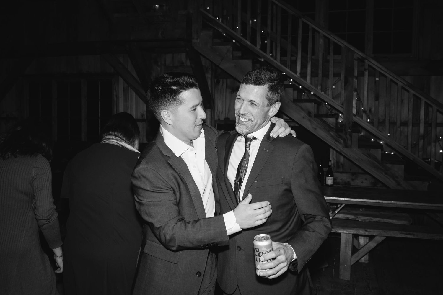 Two men in suits embrace in warm conversation at a dimly lit indoor event space, with stairs visible in the background.