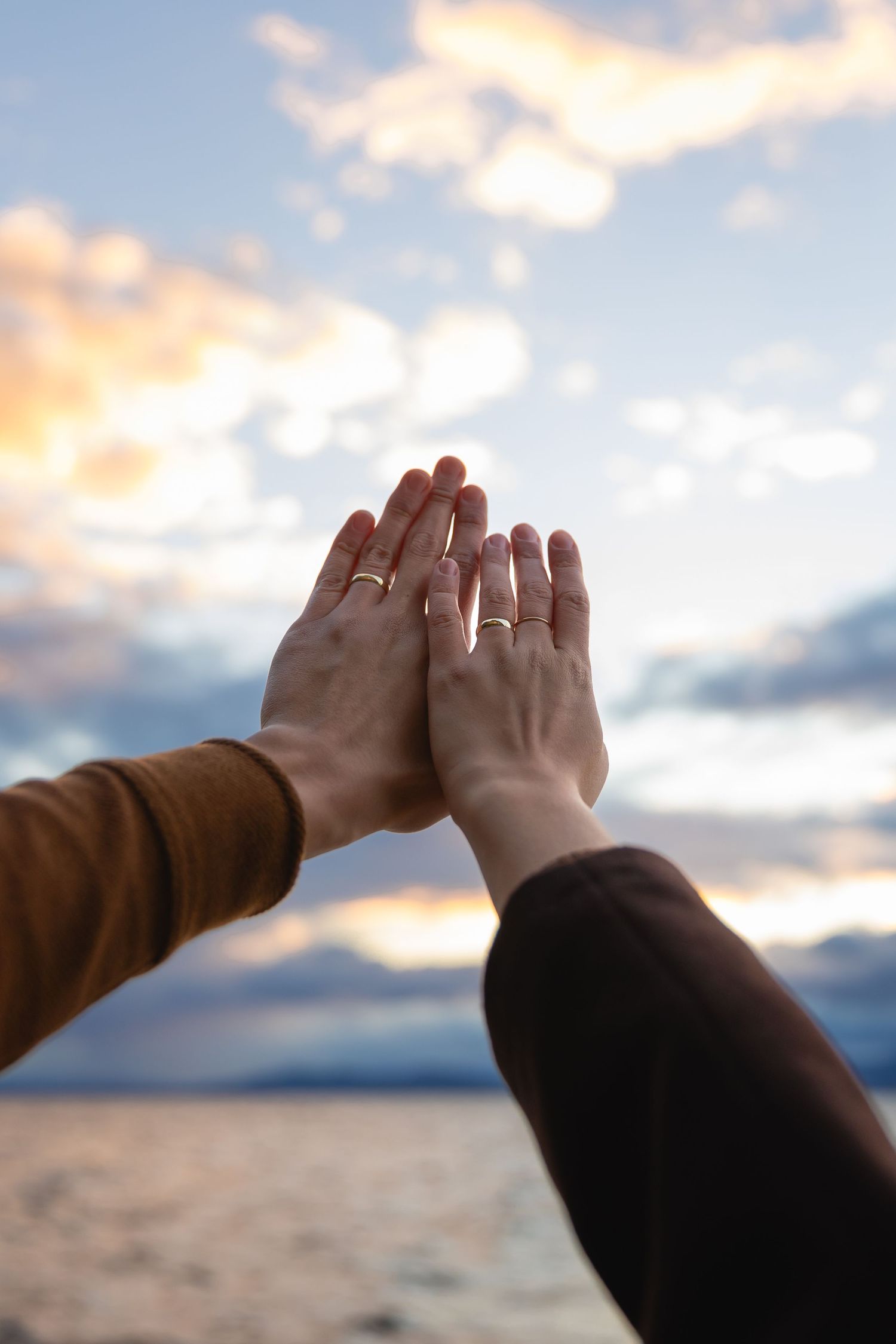 Two hands wearing rings reach up towards a dramatic sunset sky over water, creating a silhouetted high-five gesture.