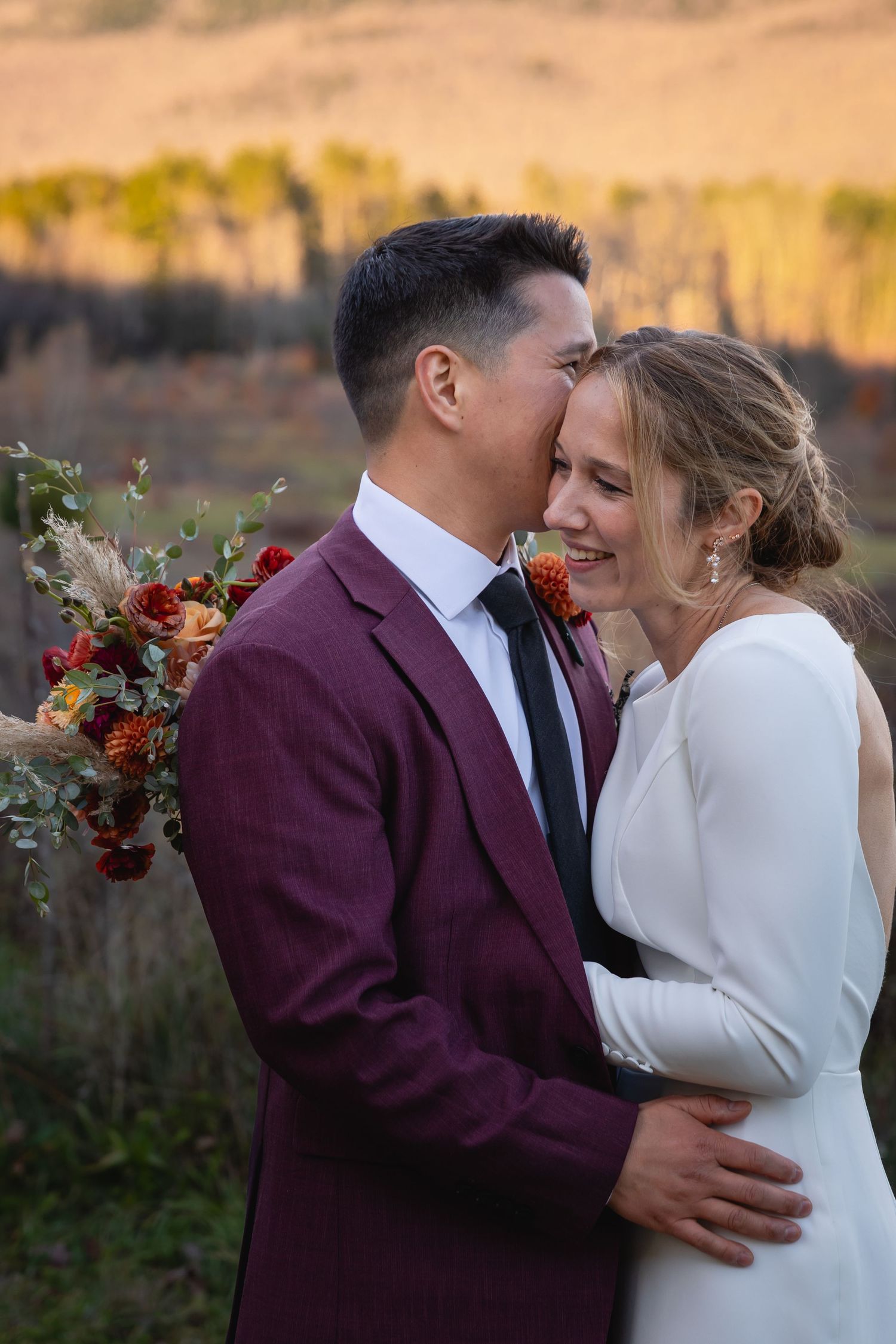 A loving embrace between newlyweds outdoors at sunset, with burgundy and white wedding attire and a colorful bouquet.