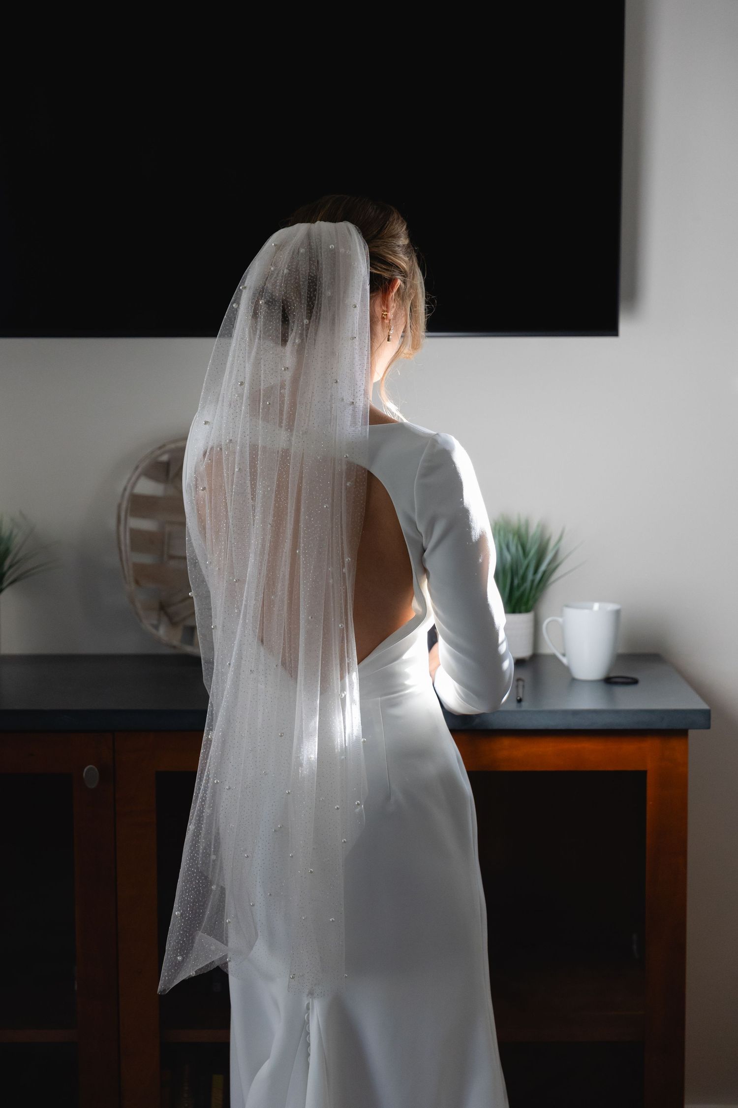 Back view of a bride in an elegant white dress with open back design and flowing veil standing indoors.